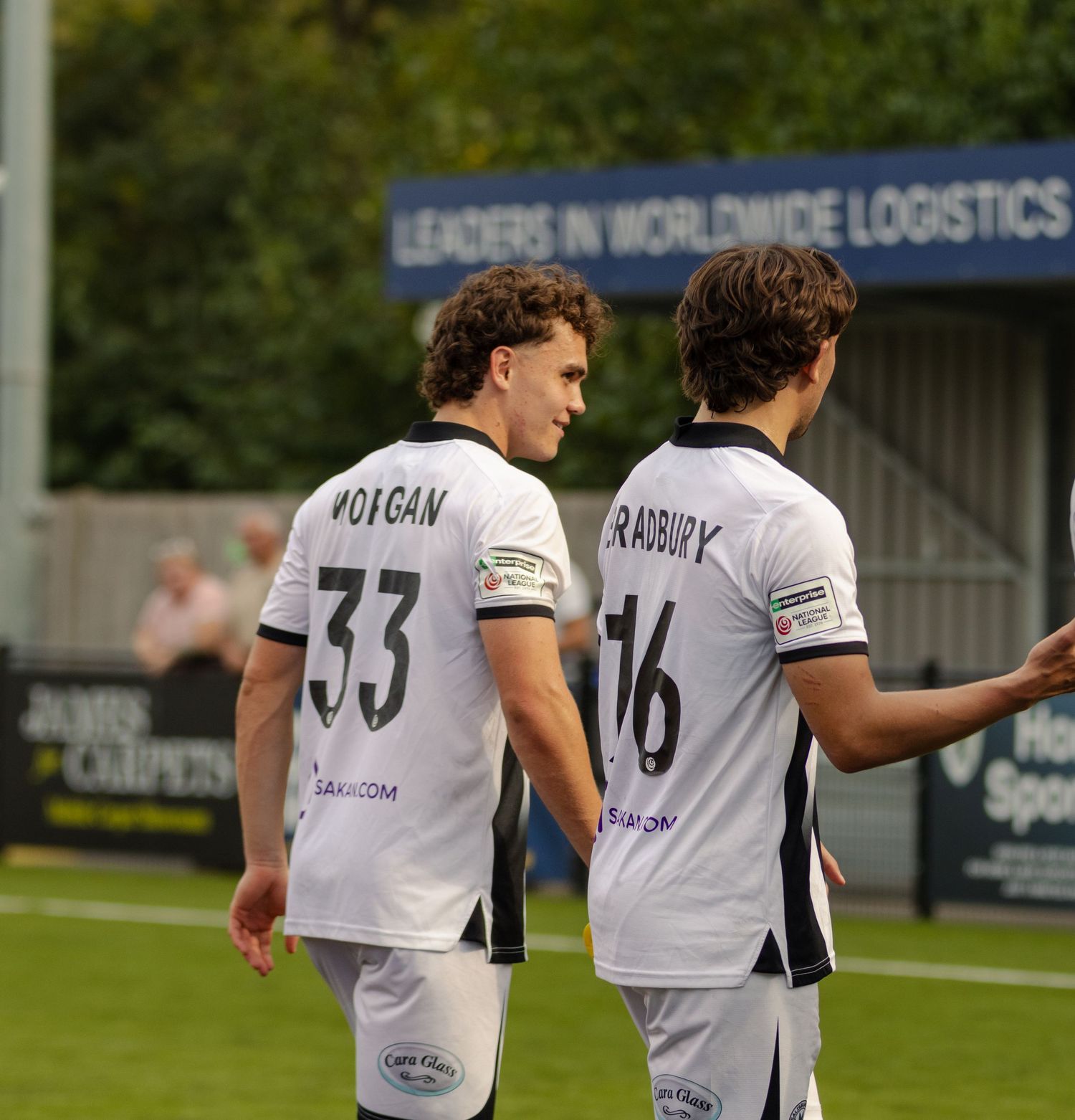 Two footballers wearing MORGAN 33 and RADBURY 16 shirts walk together on a football pitch.