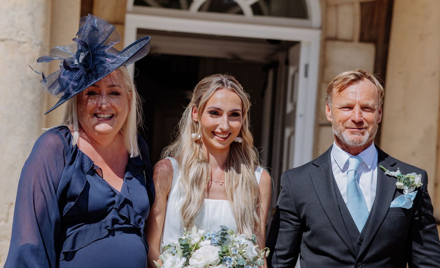 Bride poses with parents in navy blue outfits outside a stone building on her wedding day.