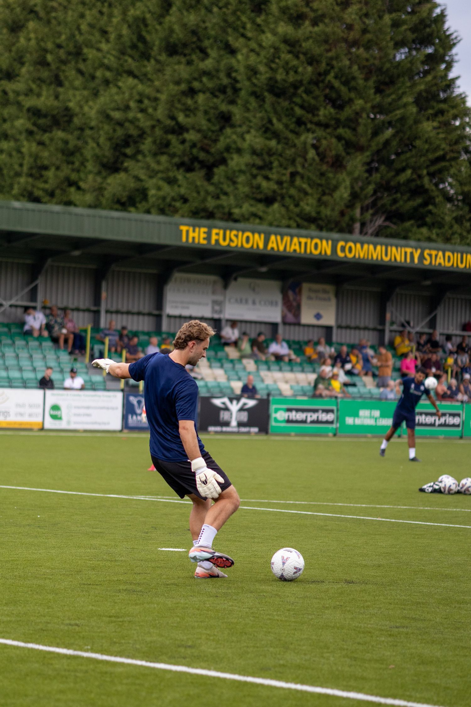Goalkeeper in blue kit kicking football during pre-match warm-up at Fusion Aviation Community Stadium.