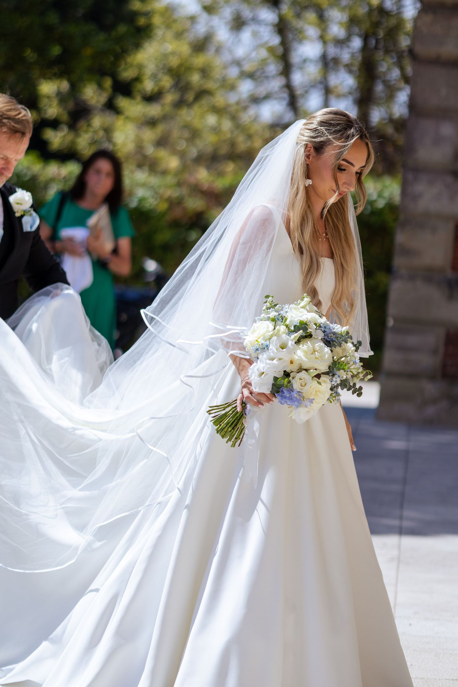 Bride in flowing white ball gown and veil walking with white and blue floral bouquet.