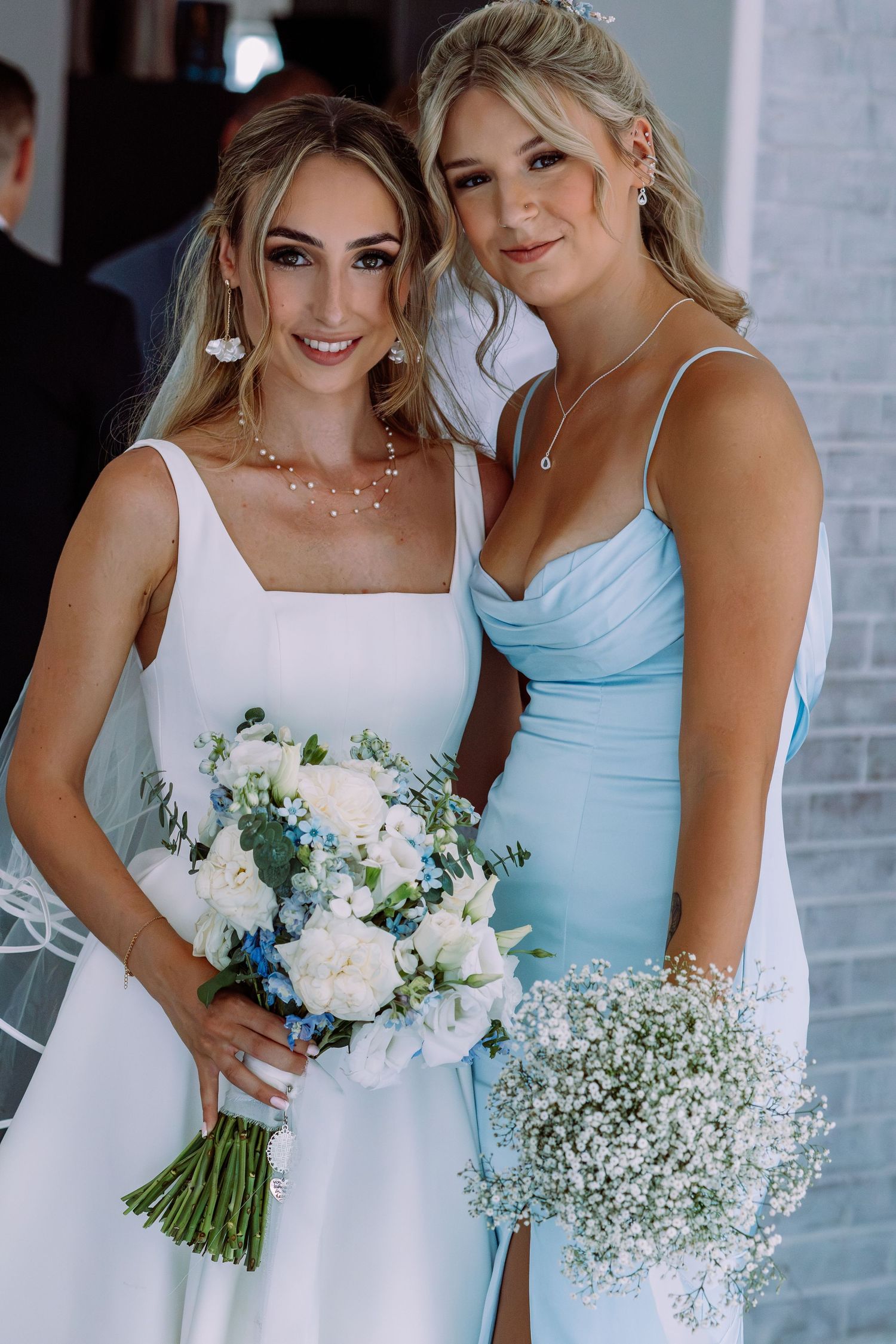 Bride in white square-neck gown and bridesmaid in light blue dress posing with floral bouquets.