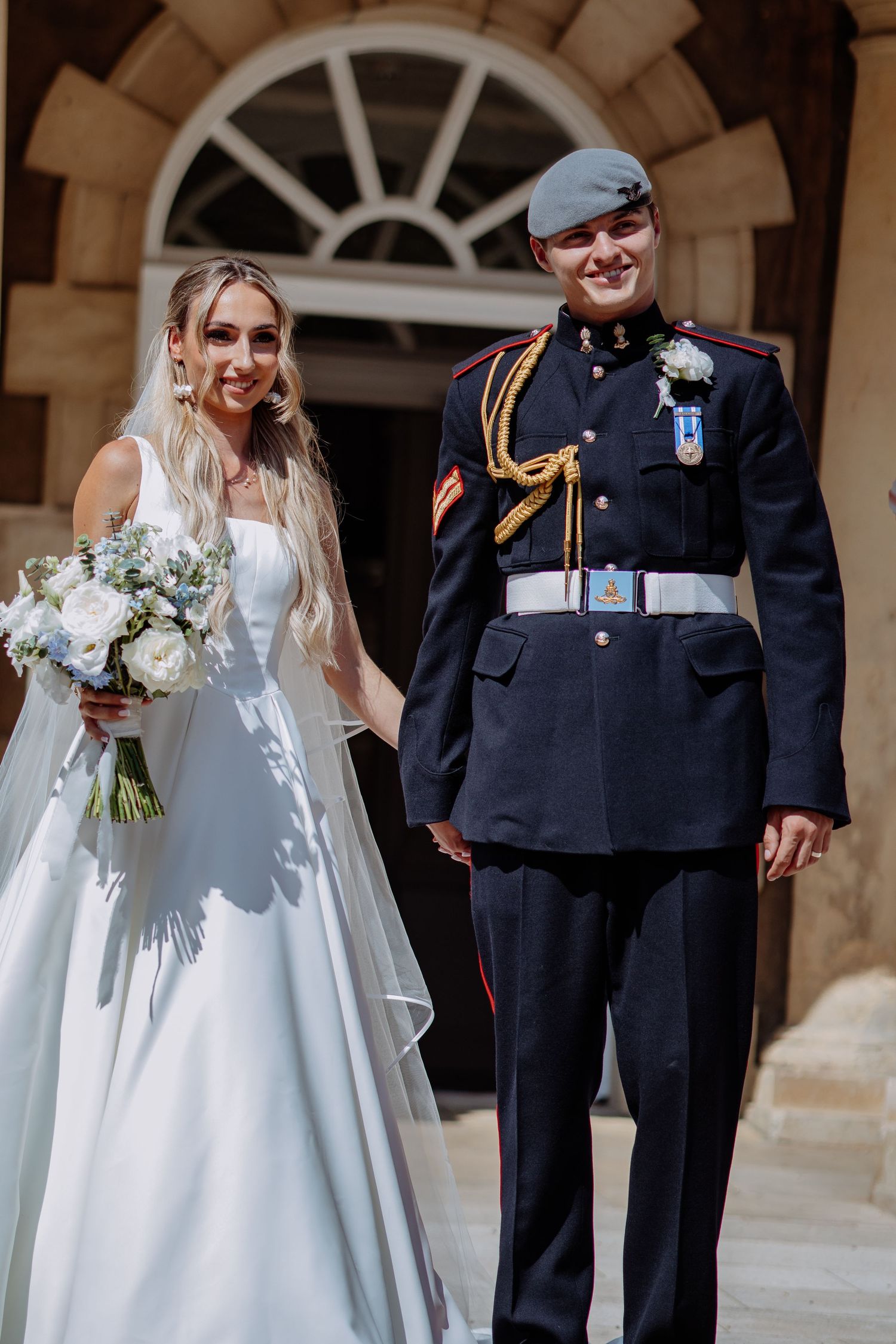 Newlyweds smile joyfully outside stone venue, bride in white gown with veil and groom in RAF military dress uniform.