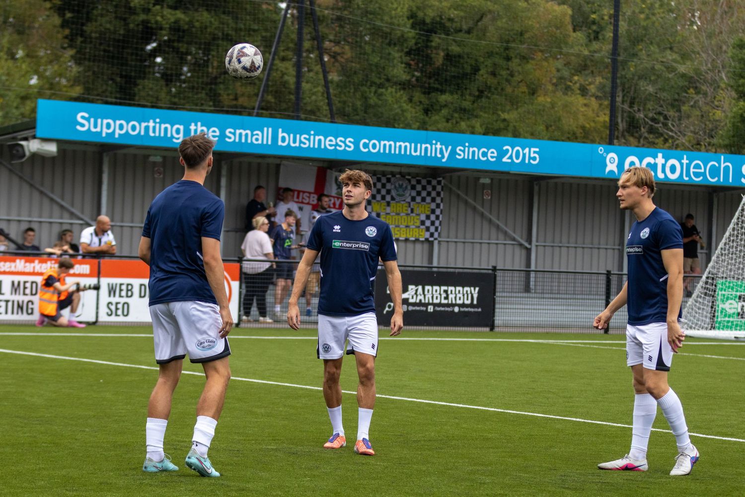 Three footballers in navy kit warming up with a ball at a non-league ground with sponsor banners visible.