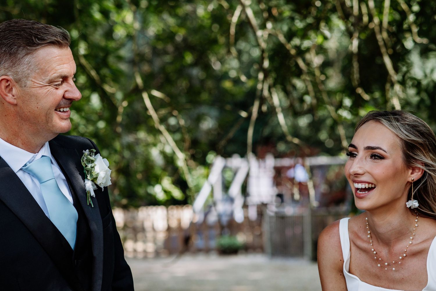 Candid outdoor wedding moment of man in black tuxedo with blue tie and laughing bride in white dress.