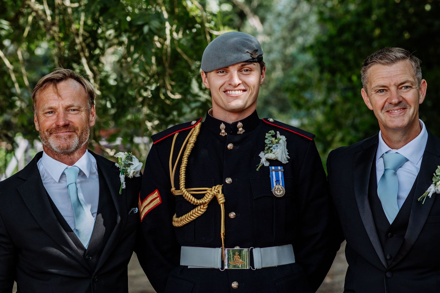 Three smiling men at wedding, center man in military dress uniform flanked by two in black suits.