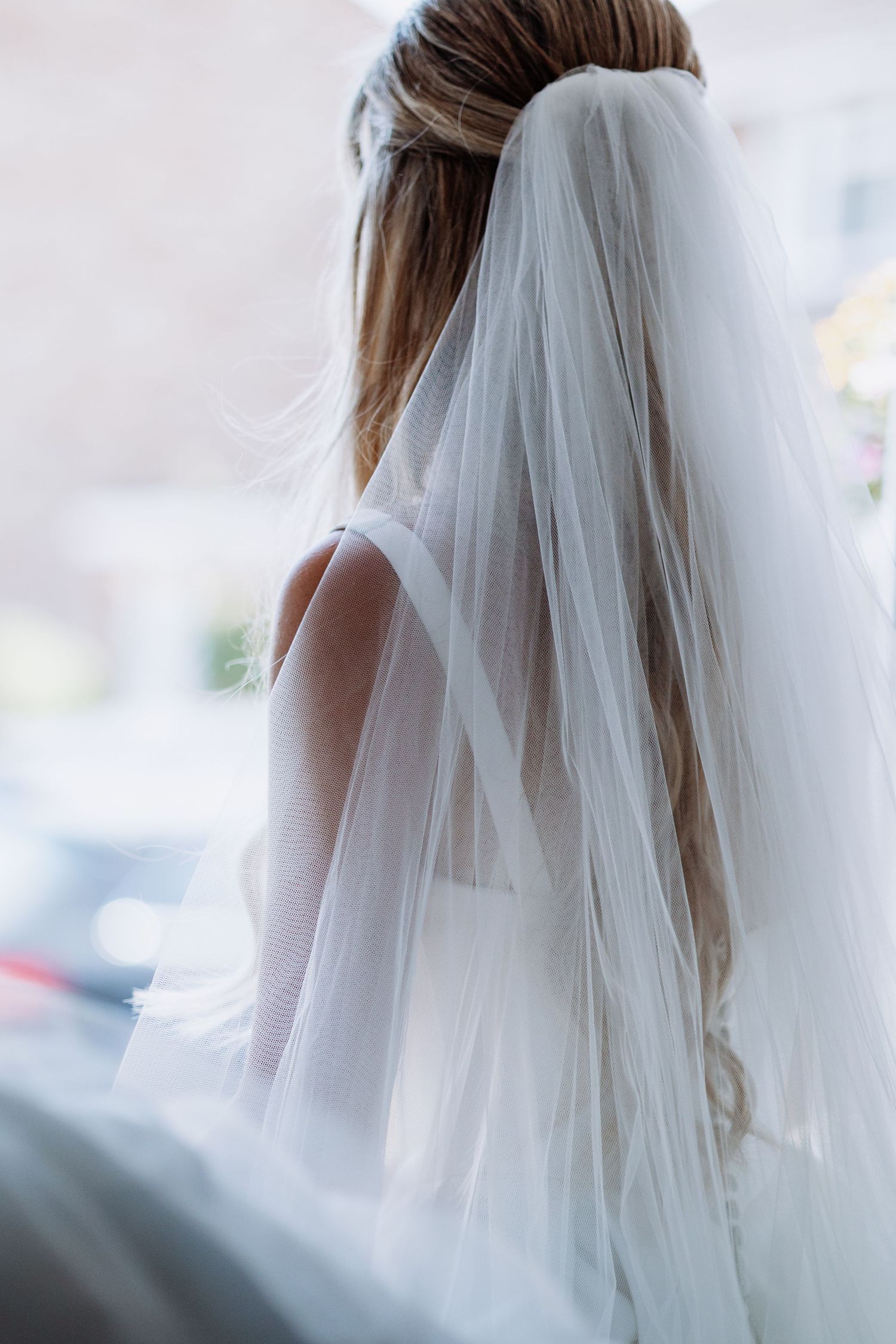 Bride wearing a white wedding veil seen from behind, long hair flowing down her back near a bright window.
