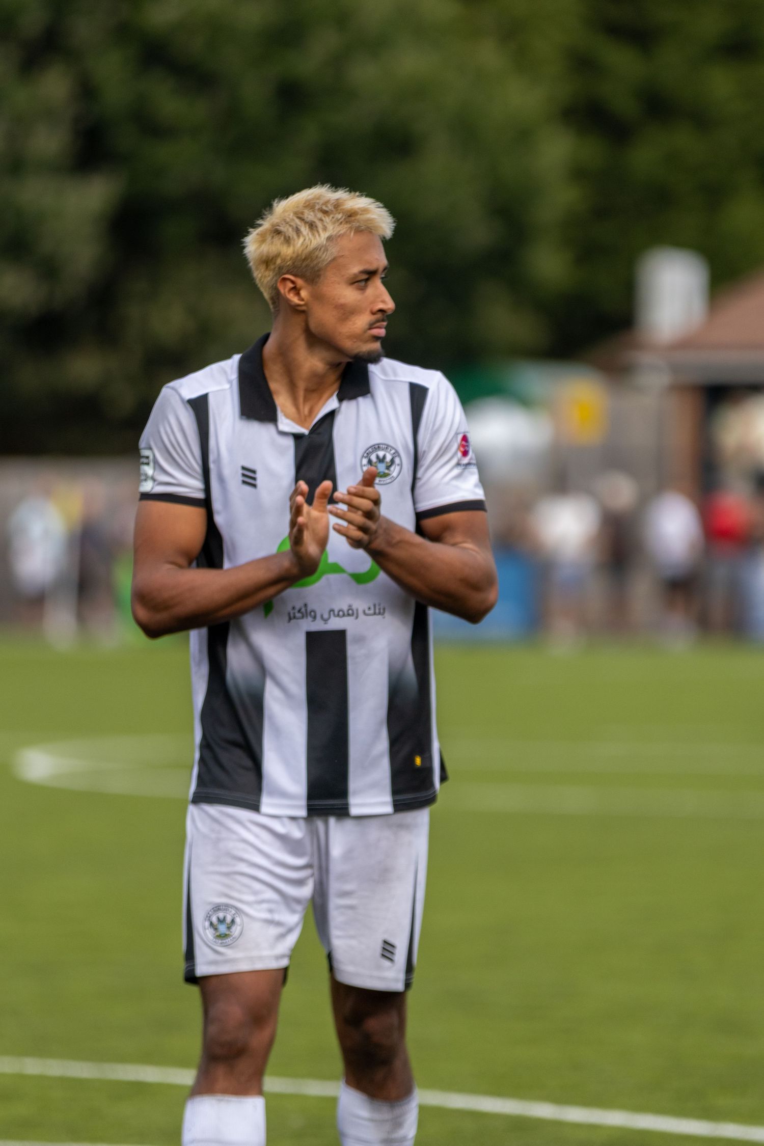 Blonde male soccer player in black and white striped jersey claps hands on a green football pitch.