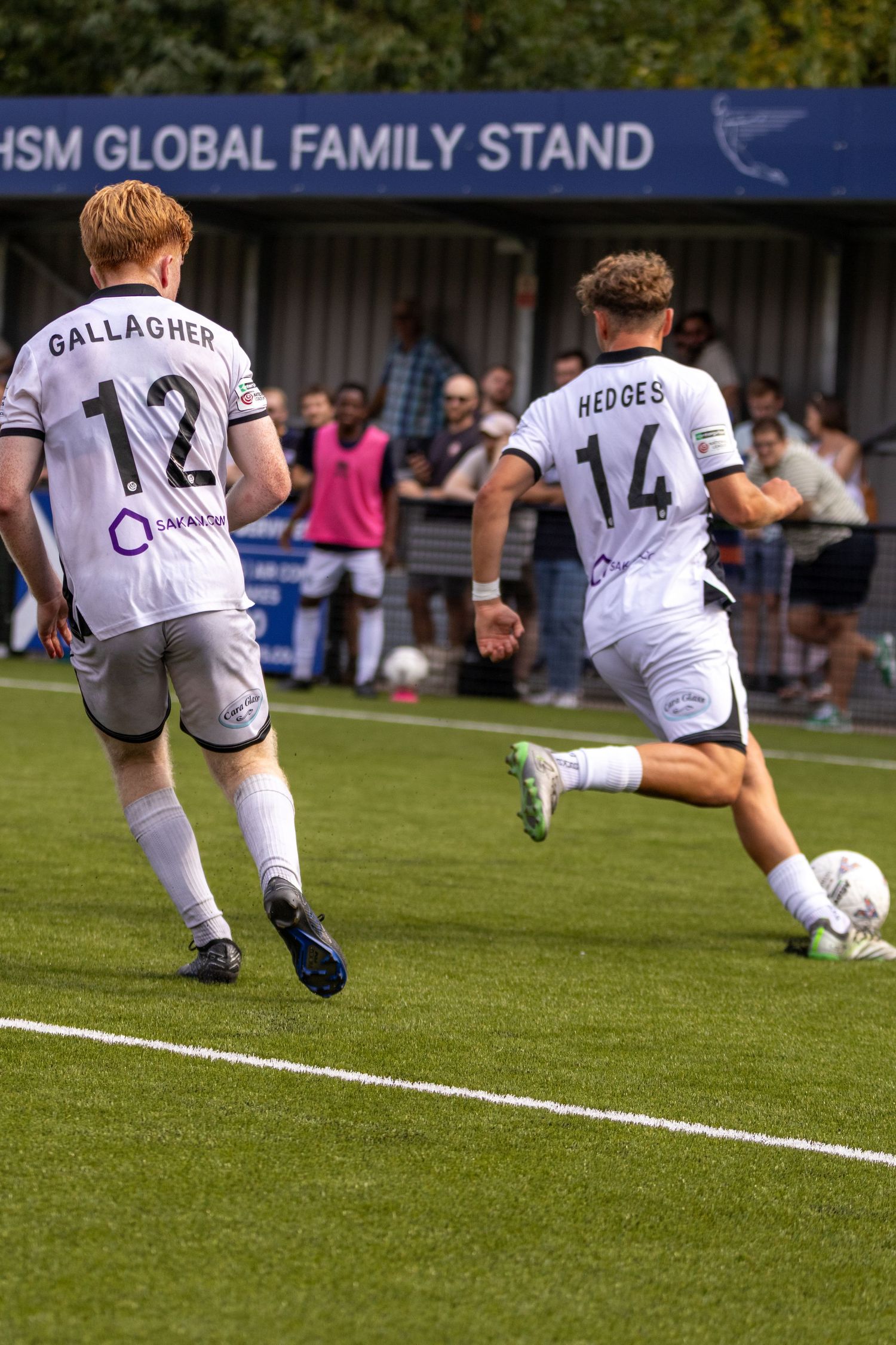 Hedges #14 strikes the ball as Gallagher #12 watches near the HSM Global Family Stand in a football match.