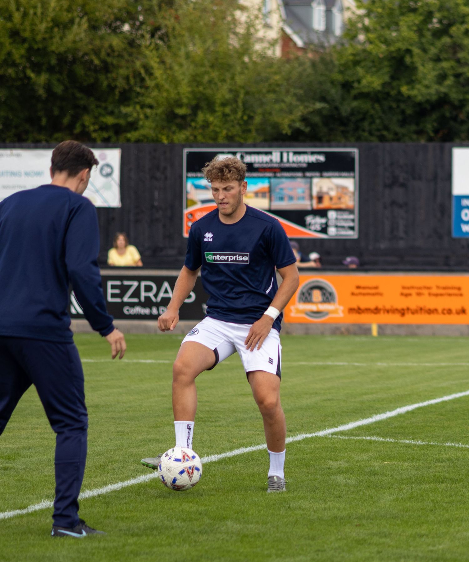 Young male soccer player in navy blue kit controlling a ball during pre-match warmup at a non-league ground.