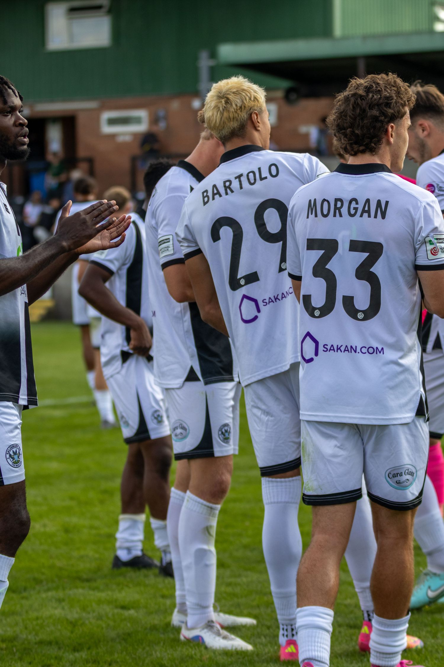 football players in white jerseys with numbers 29 Bartolo and 33 Morgan line up on a green field after a match.