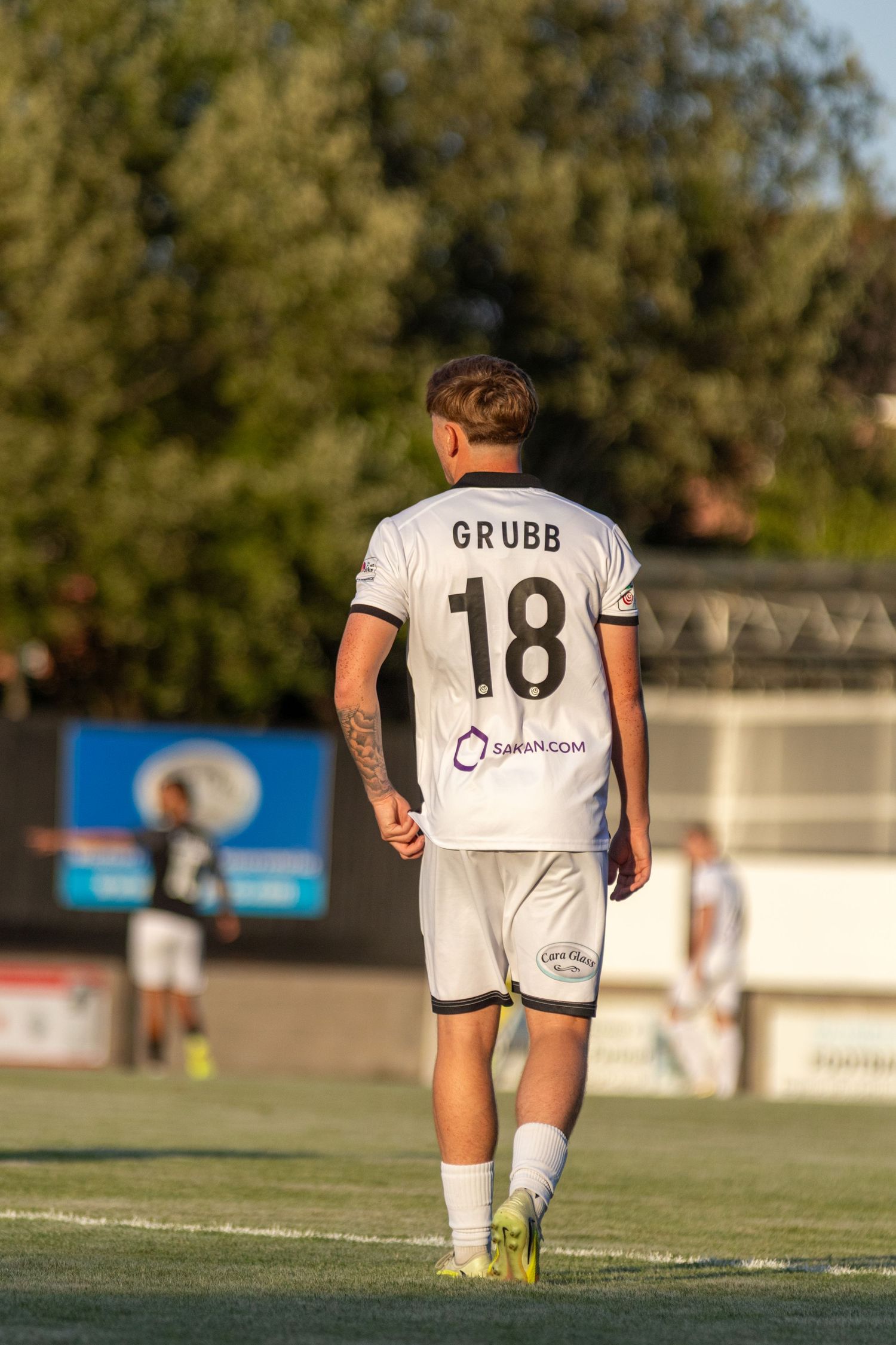Rear view of footballer Grubb number 18 in white kit with Sakan.com sponsorship walking on pitch.