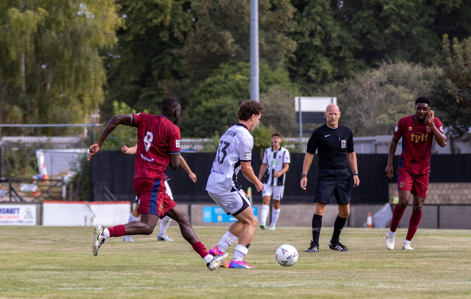 Salisbury FC midfielder drives forward with the ball as referee watches during a sunny fixture.