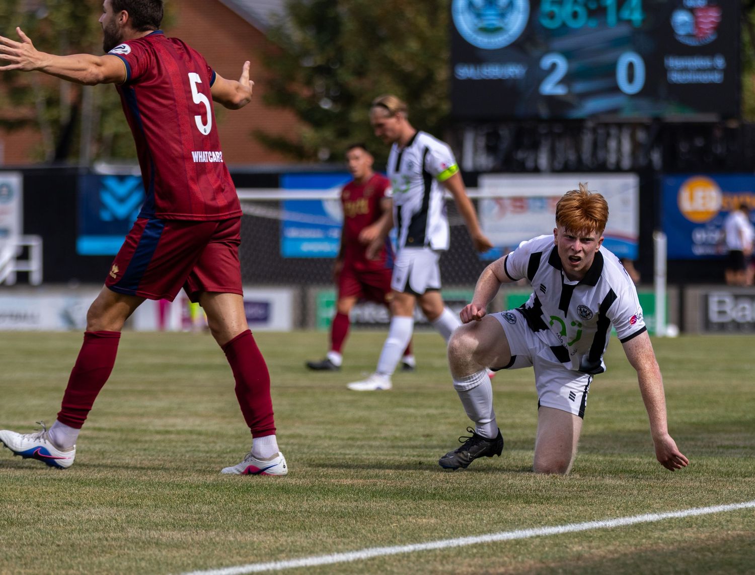 A red-haired footballer in black and white kit kneels on the pitch during a 2-0 match.