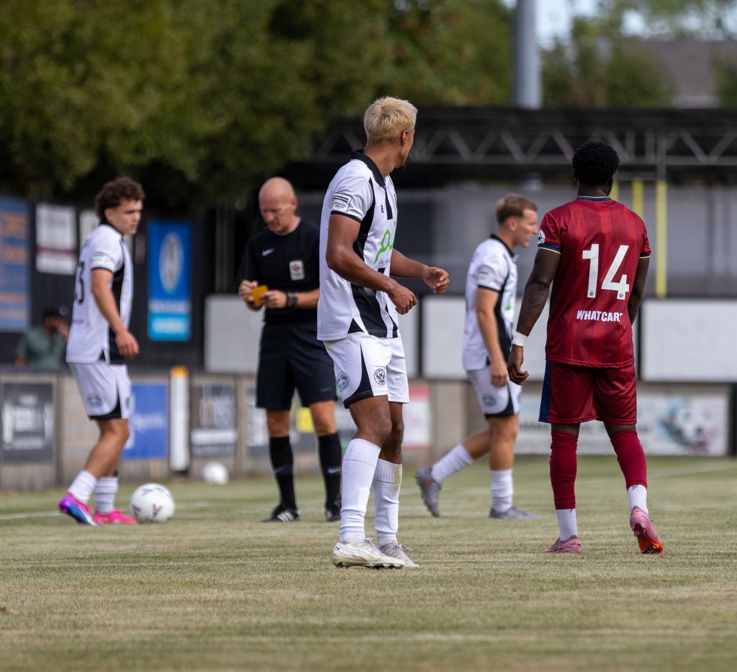 Football players in black and white kit face opposition in red during an outdoor match with referee.