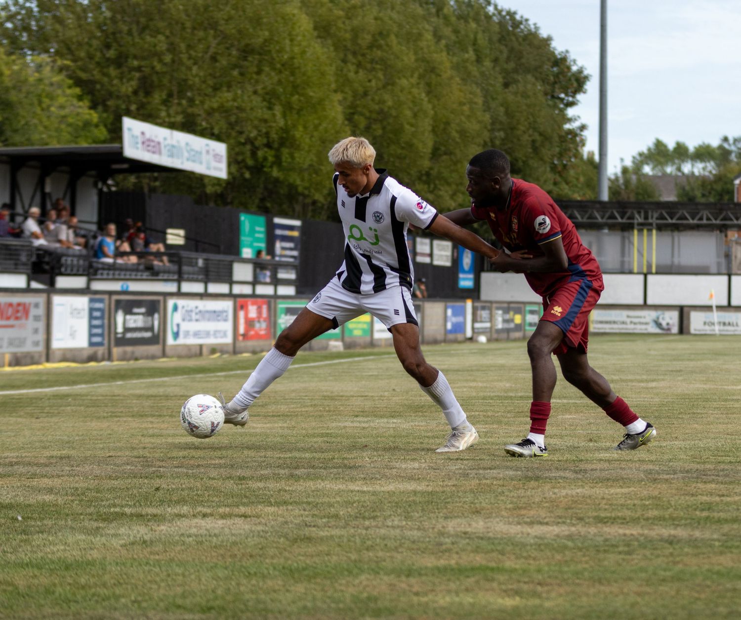 A footballer in black and white stripes dribbles past a red-kitted opponent during an outdoor football match.