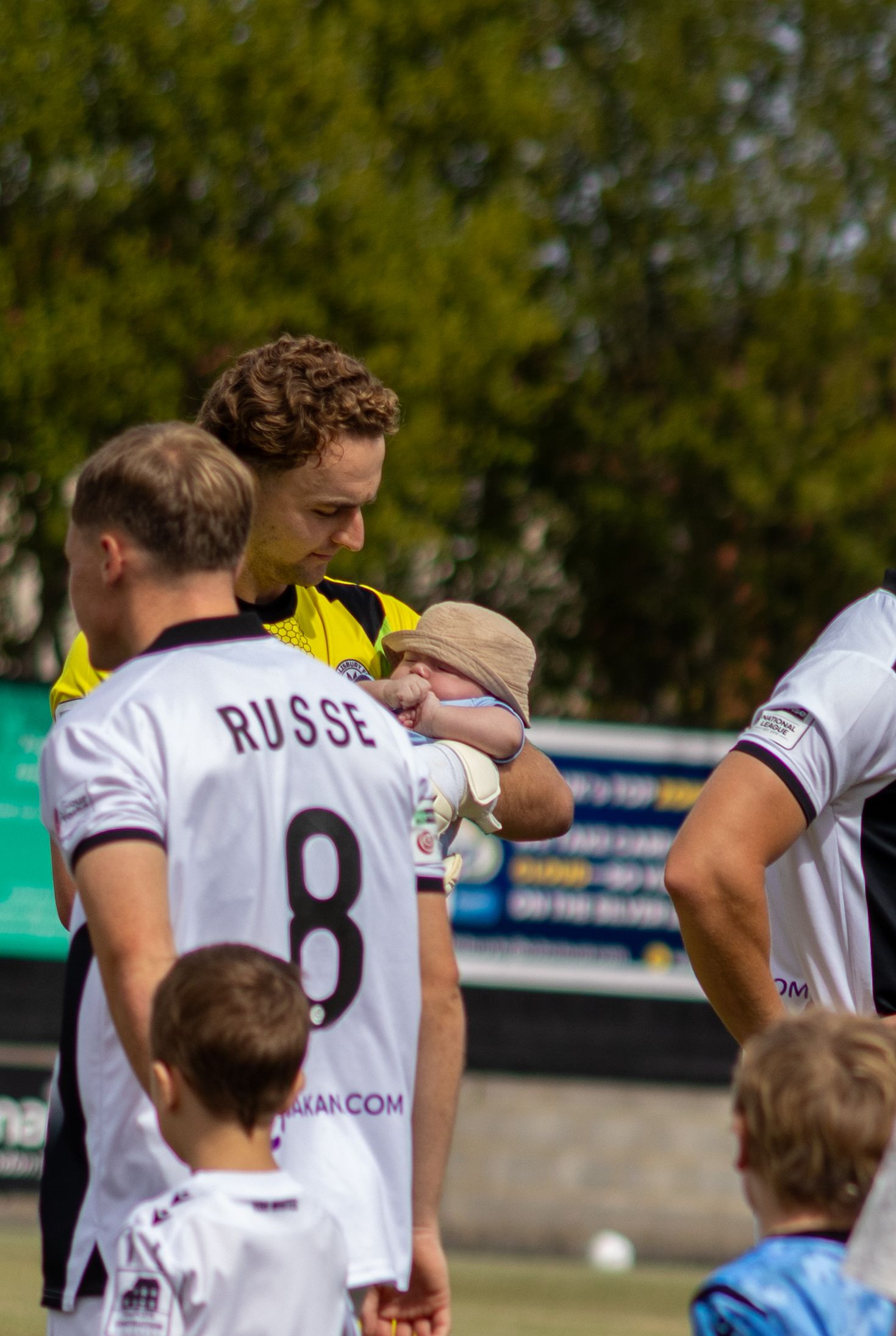 A football referee in yellow kit holds a baby while standing with players in white jerseys before a match.