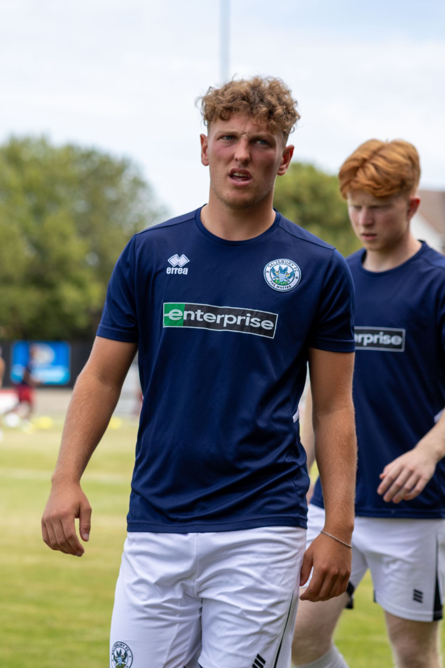 Curly-haired footballer in navy Enterprise-sponsored shirt walking on pitch during pre-match warmup session.