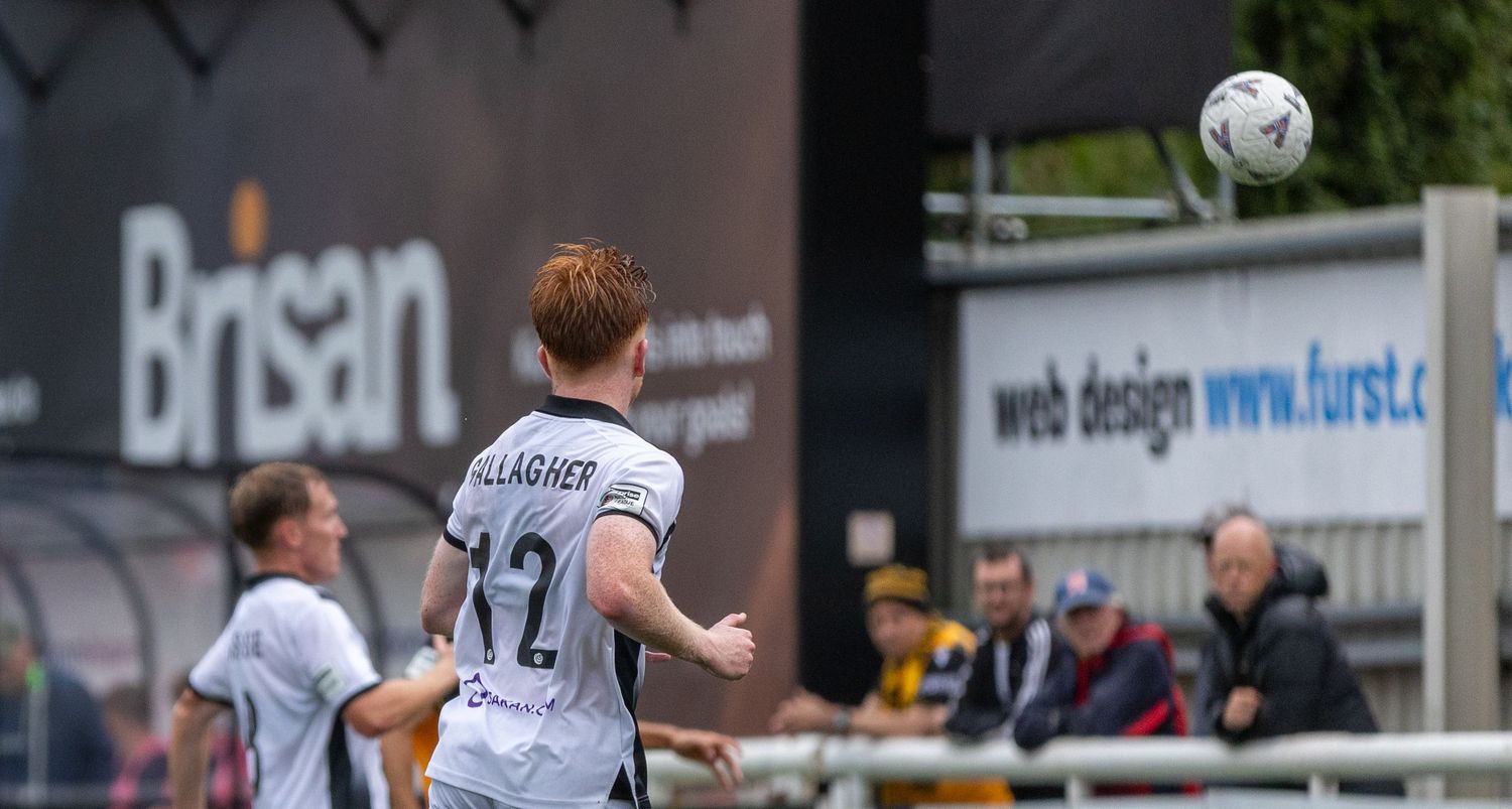 A red-haired footballer in white kit heads the ball during an outdoor match near advertising boards.