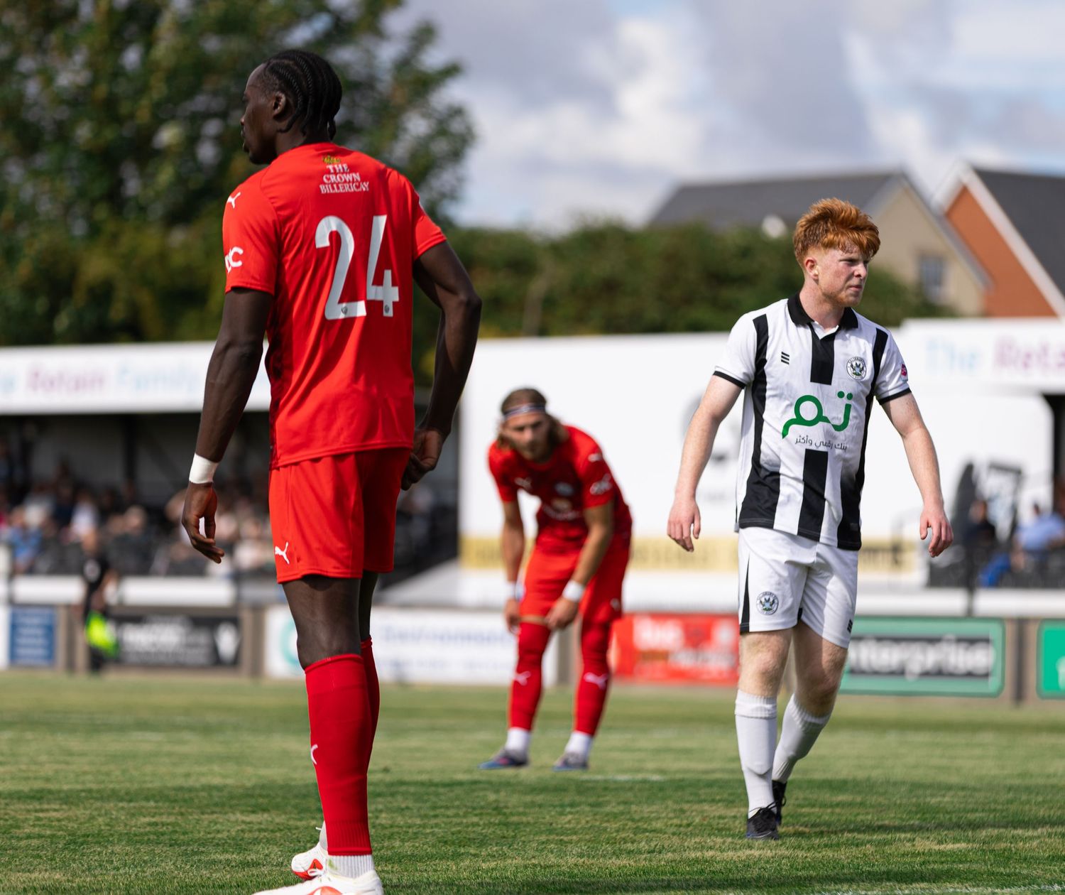 Football players in red and black-and-white striped kits compete on a sunny pitch, with number 24 in red prominent.