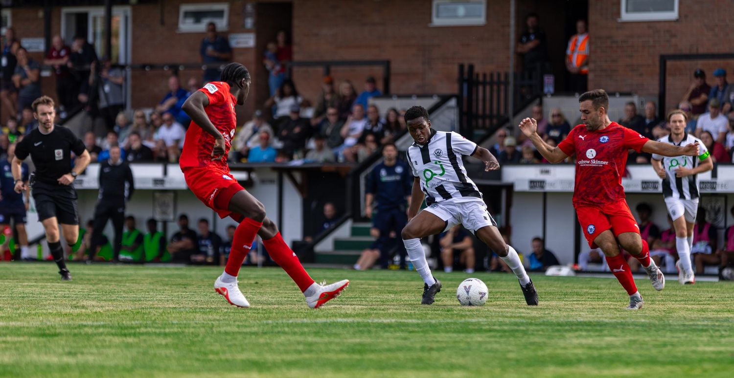 Soccer player in black and white kit shoots on goal while red-kitted defenders attempt to block the shot.