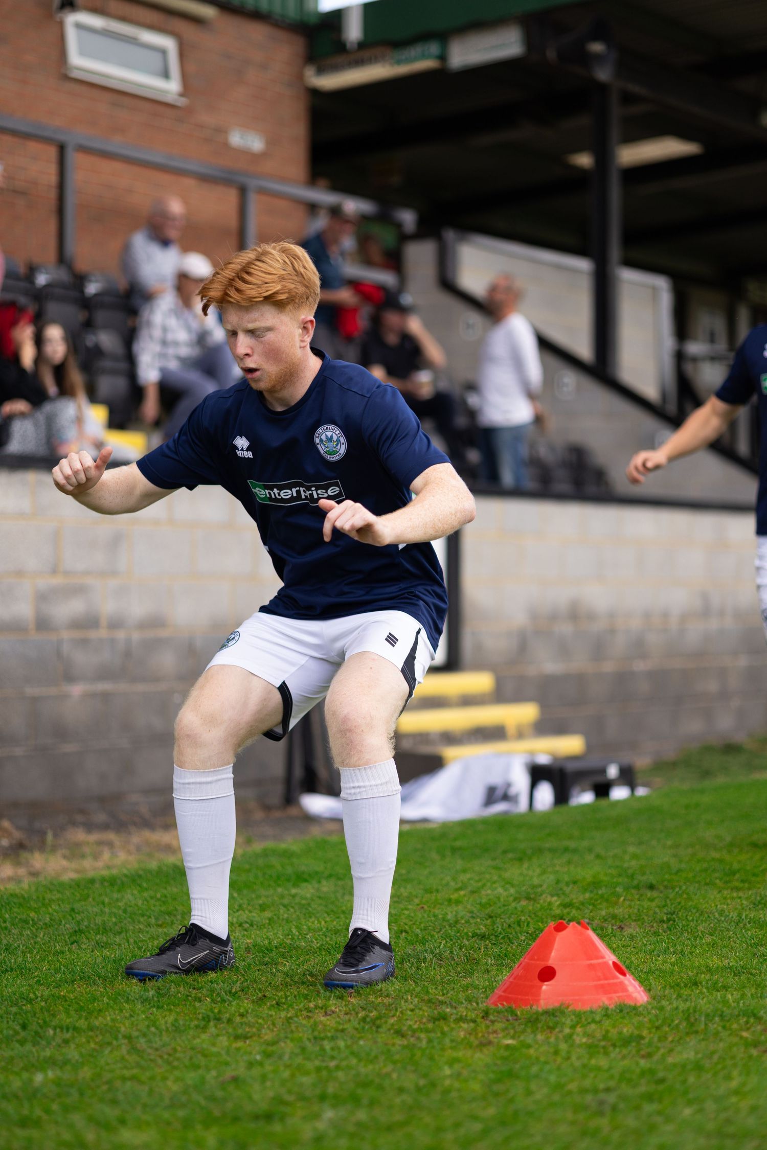 Young male soccer player in navy kit performing agility drills over orange cones on green pitch during training.