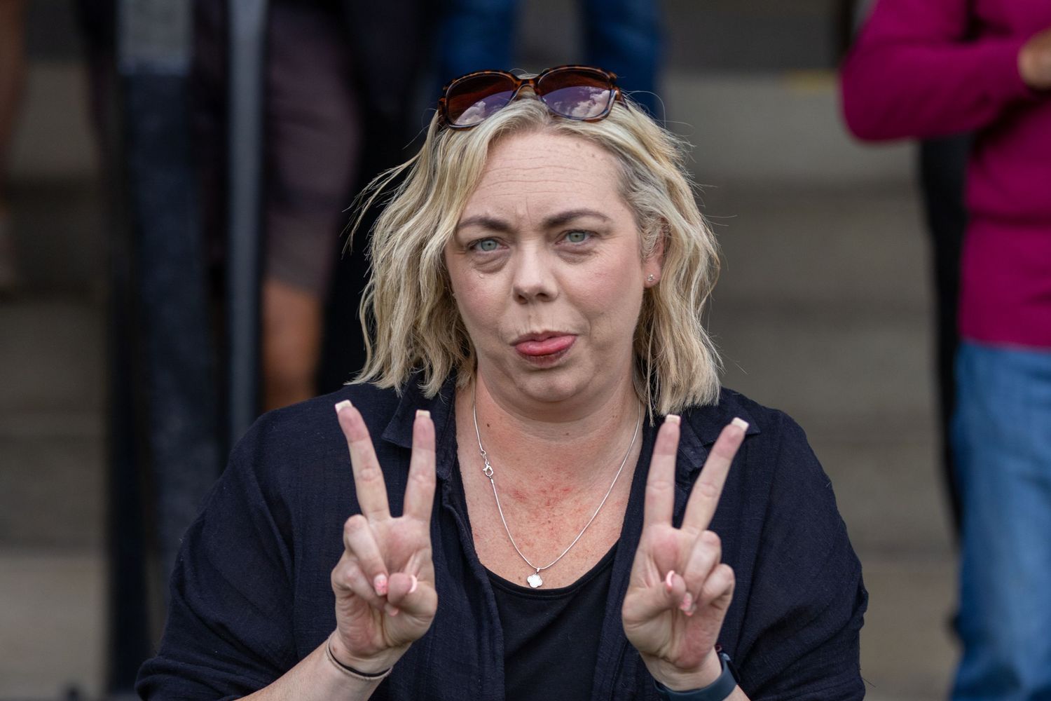 A blonde woman making double peace signs with her fingers outdoors in a black top.
