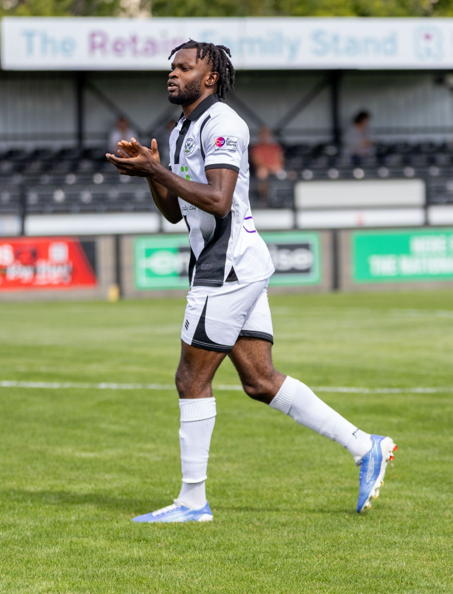 Athletic footballer in white Sakan.com kit with blue boots gesturing during a sunny match day.