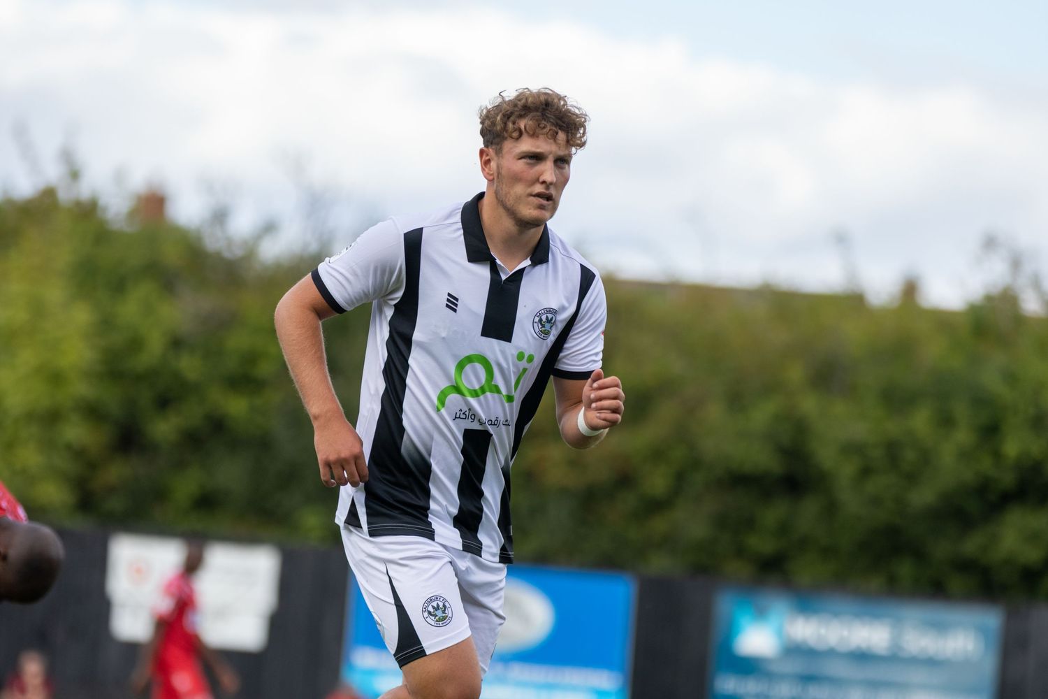 A young male soccer player in a black and white striped kit running on a green football pitch during a match.