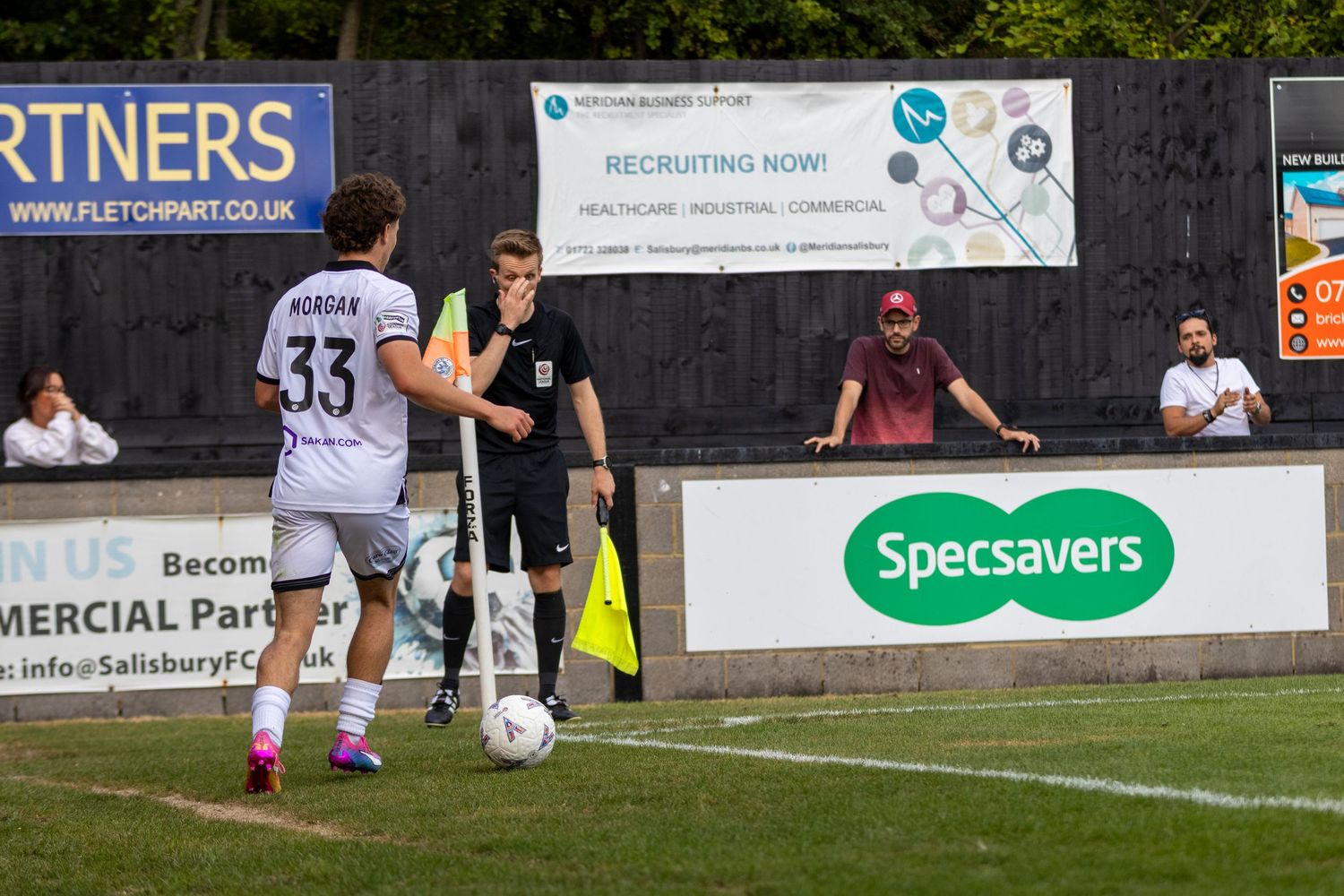 Soccer player number 33 takes a corner kick near a linesman during a match, with Specsavers advertising boards visible.