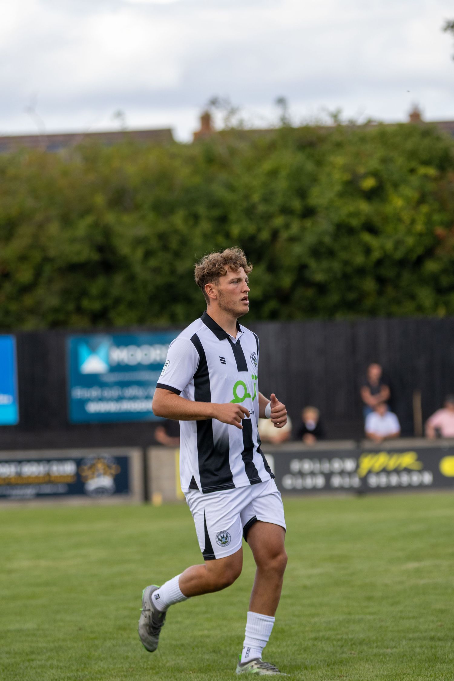 A young male soccer player in black and white striped kit jogging on a grass pitch during a match.
