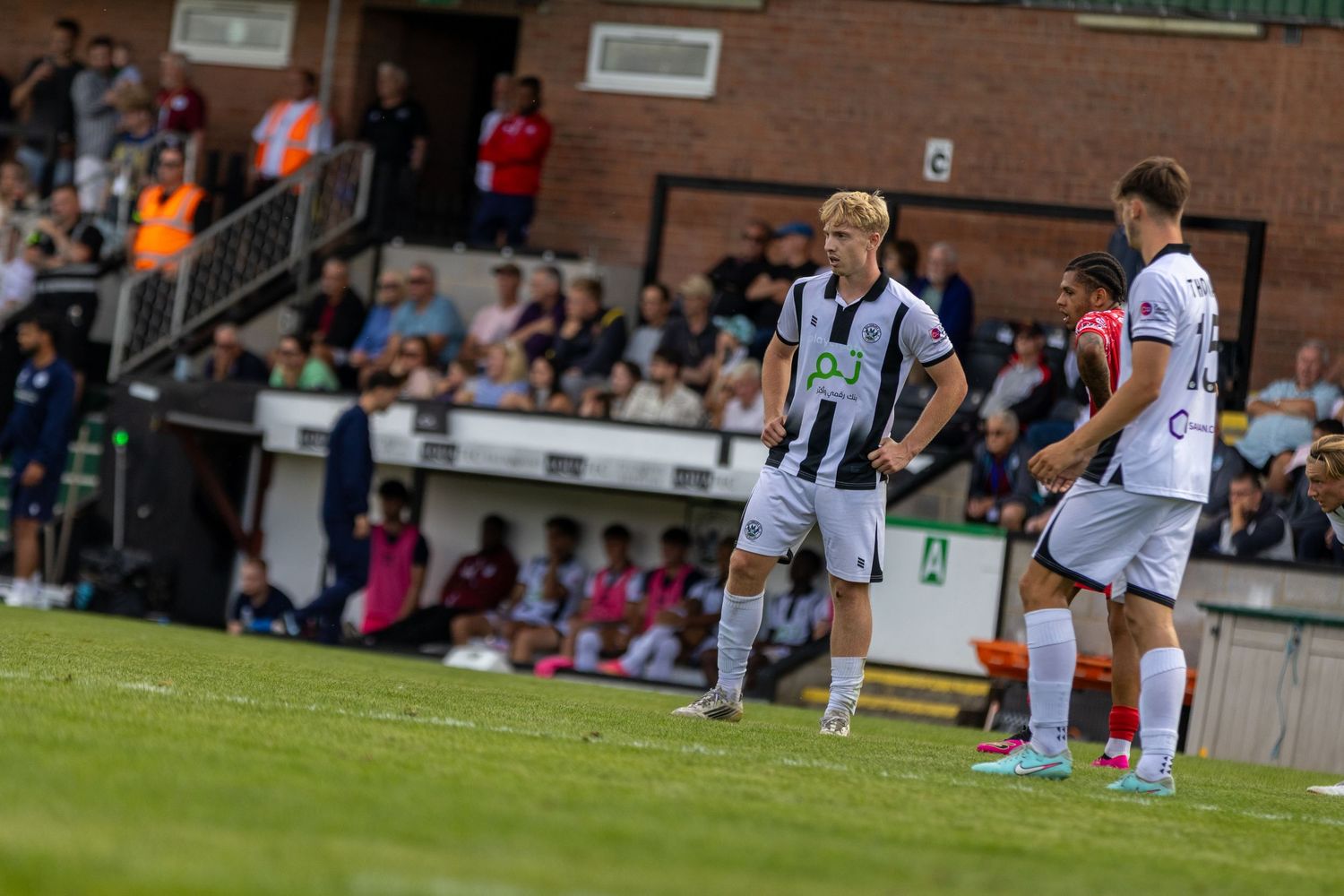 Two soccer players in black and white uniforms stand on a green pitch during a match with spectators in the background.