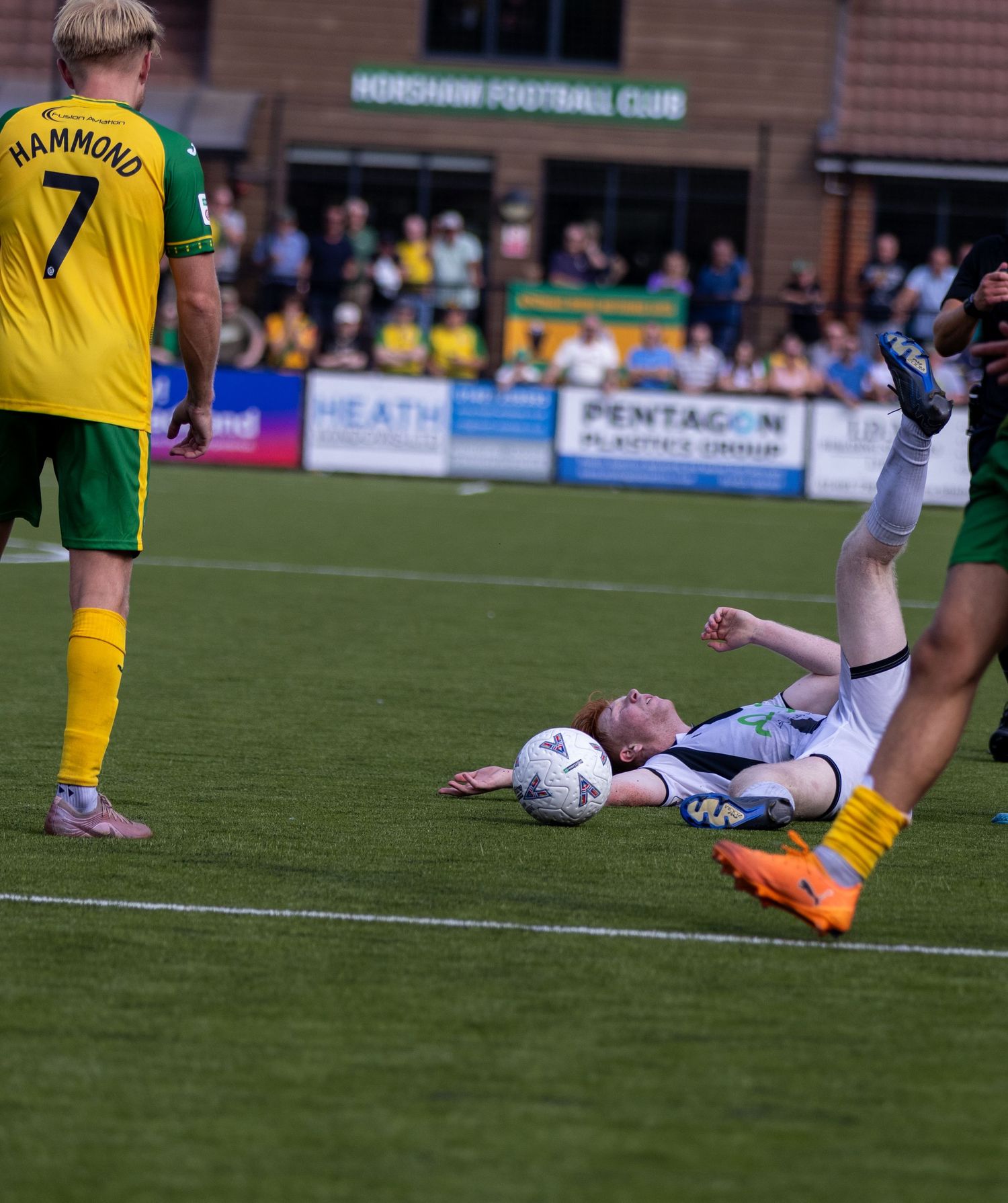 Football player in white kit slides on grass near ball as opponent Hammond number 7 watches.