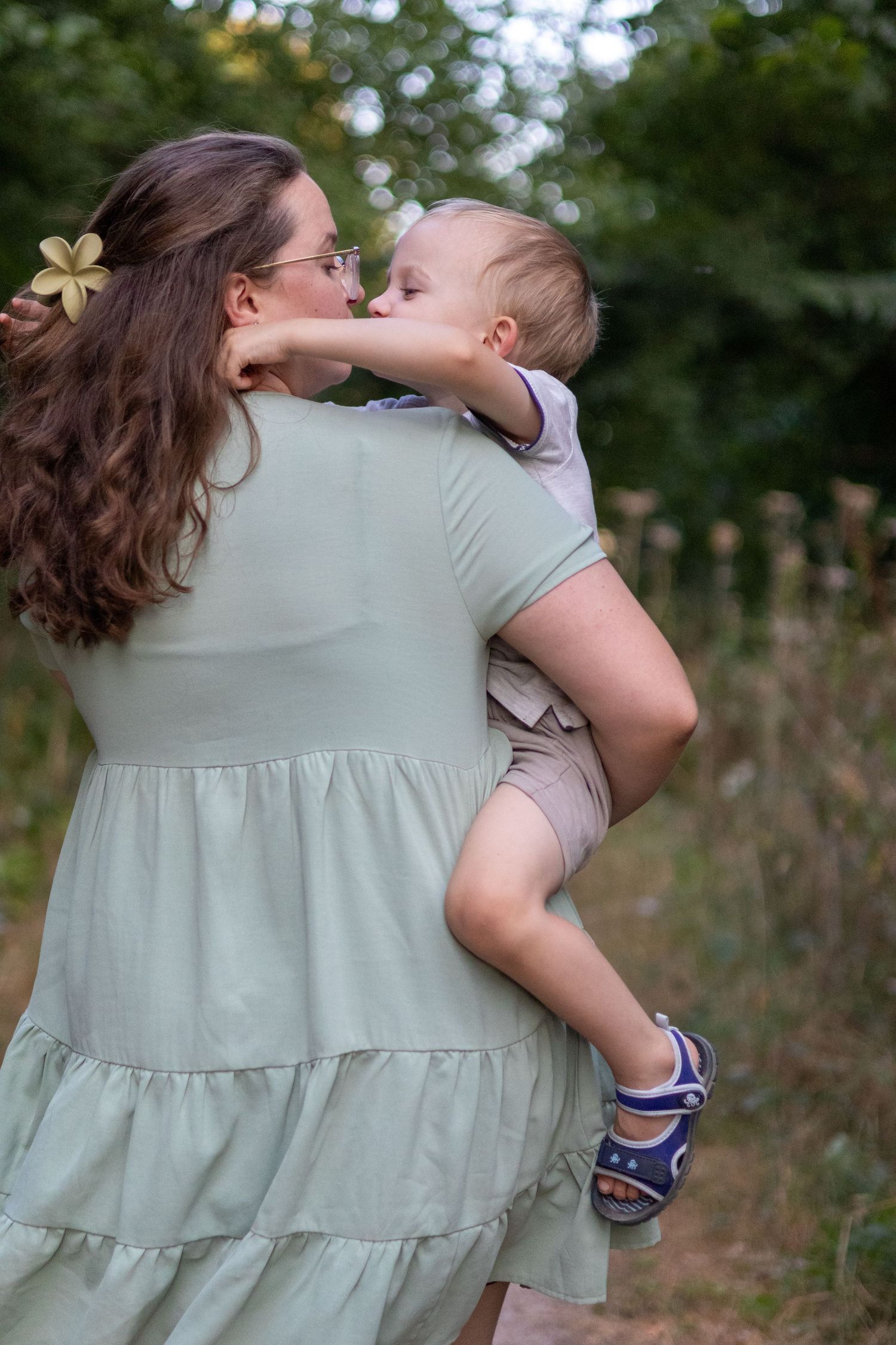 Mother holding and kissing her baby boy on a lush green nature trail outdoors.
