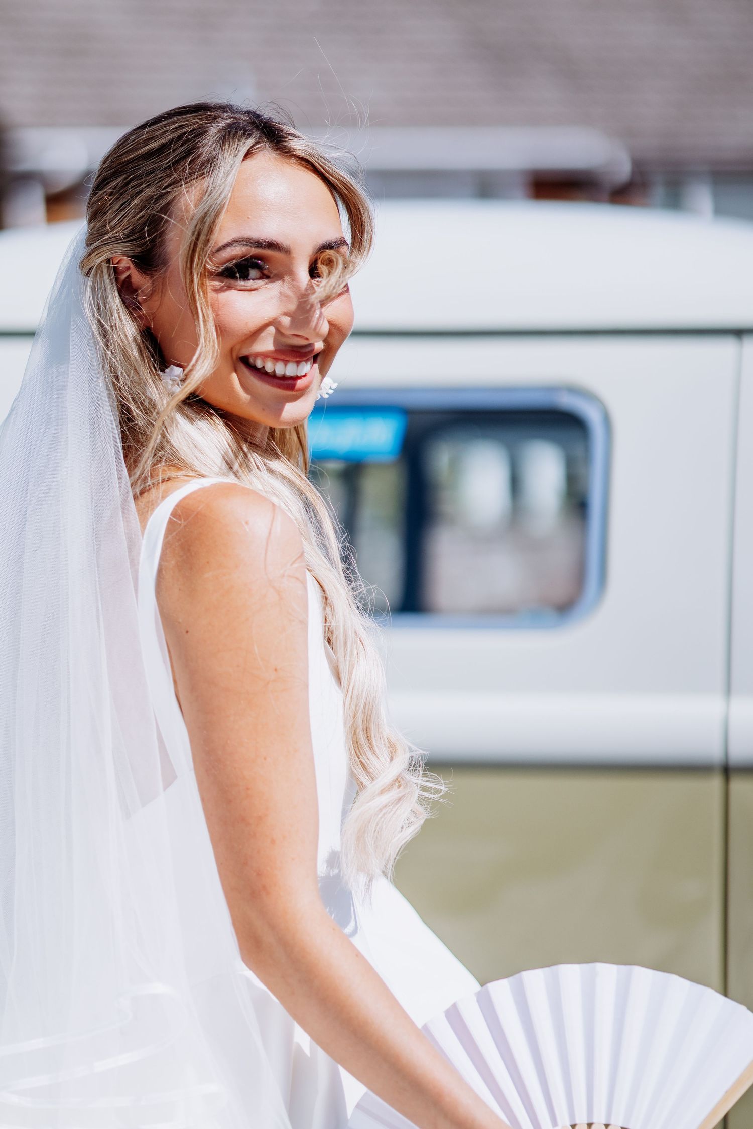 Smiling blonde bride in white wedding dress and veil holding a white fan outdoors on her wedding day.