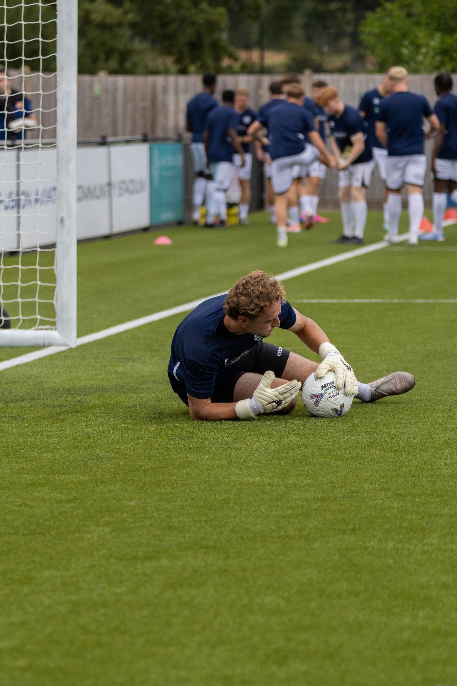 Goalkeeper in navy kit making a low save during warmup drill in front of goal with teammates in background.