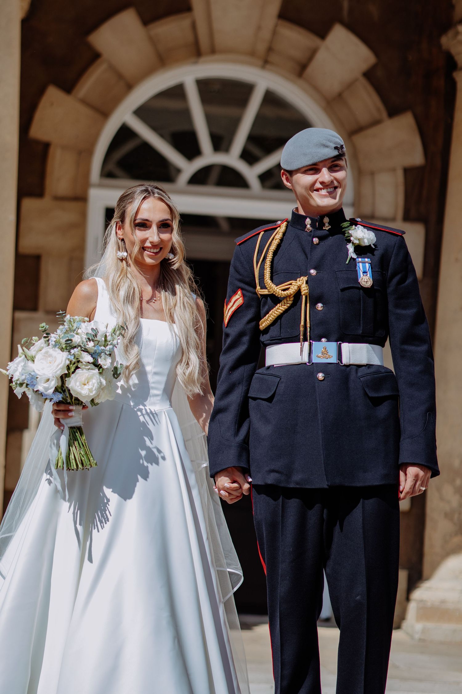 Happy couple exits ceremony venue, bride in white satin gown holding white bouquet and groom in RAF dress uniform.