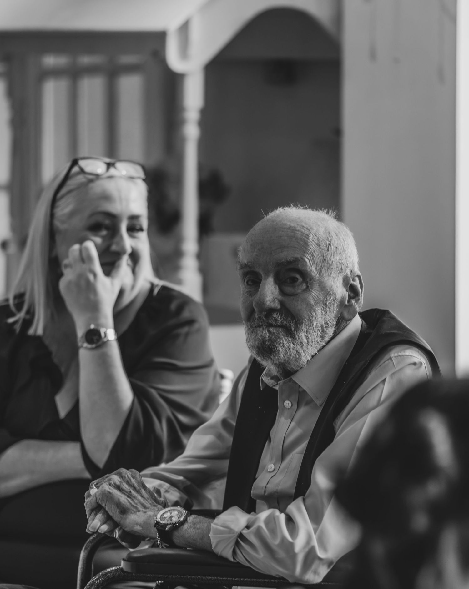Black and white candid portrait of elderly bearded man and blonde woman with glasses at indoor gathering.