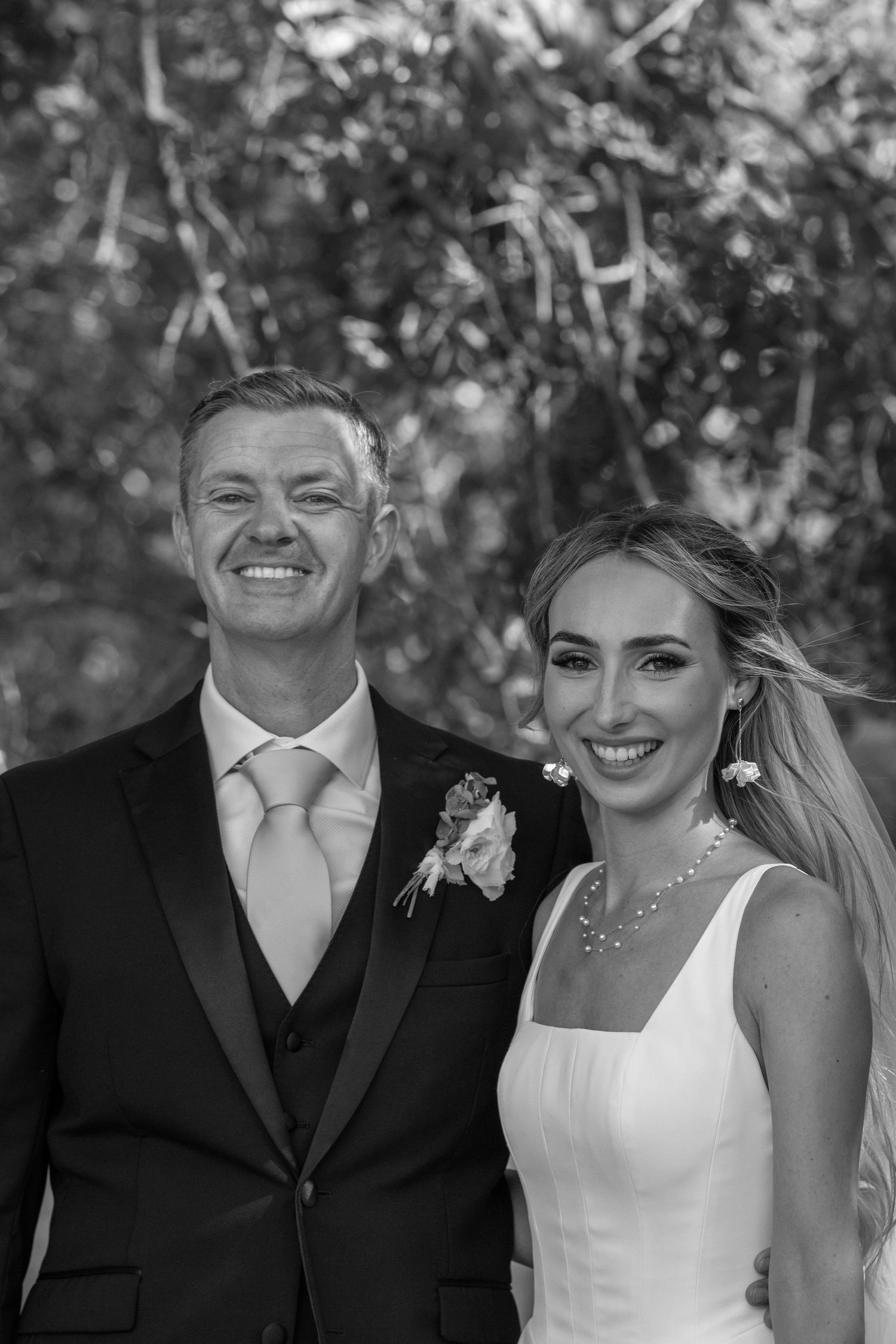 Black and white portrait of smiling groom in tuxedo with pink tie beside bride in white wedding gown.