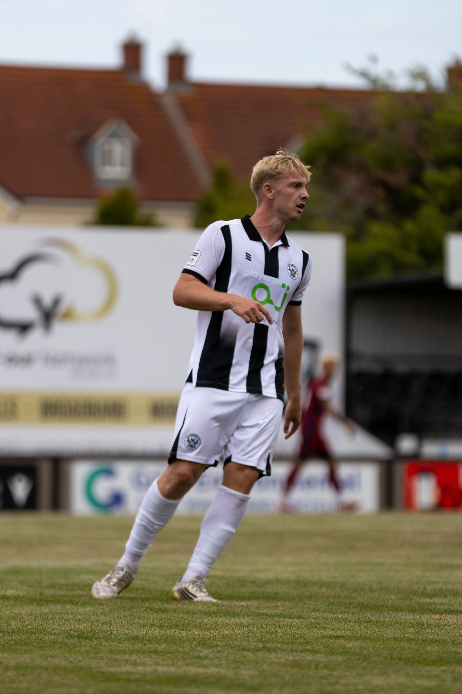 A blonde male footballer wearing a black and white striped kit jogs on a grass pitch.