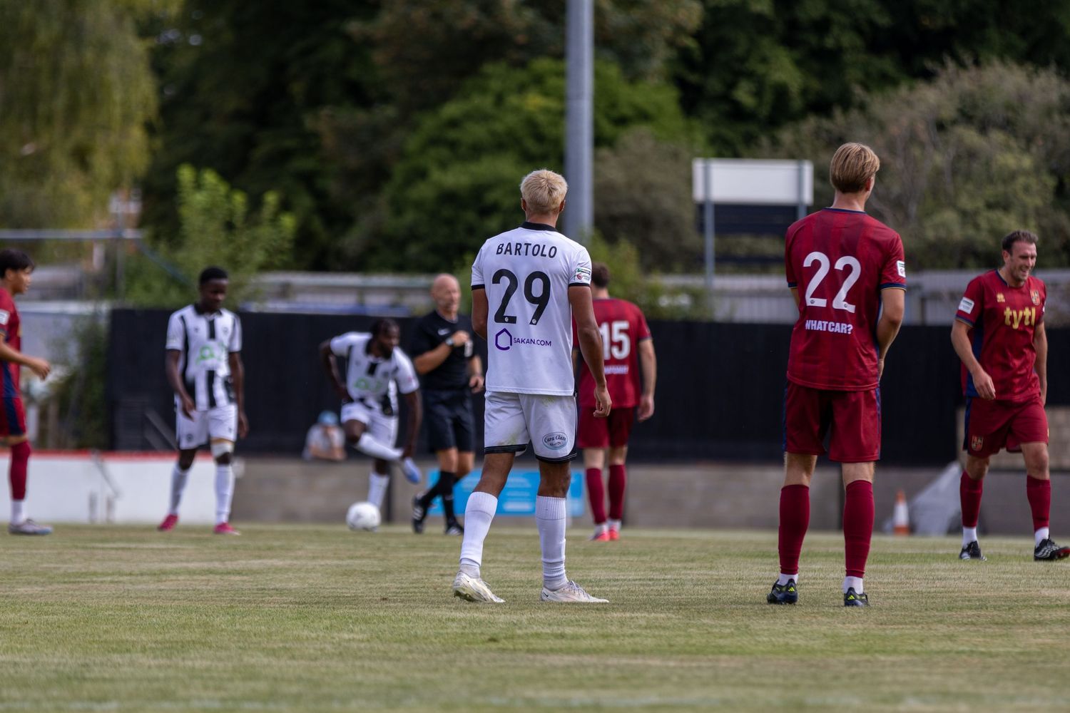 Soccer player number 29 Bartolo in white kit stands on green pitch during competitive match with red-shirted opponents.