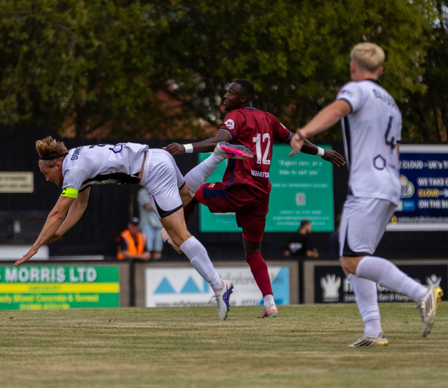 A footballer in claret kit challenges an opponent with a high kick during a competitive outdoor match.