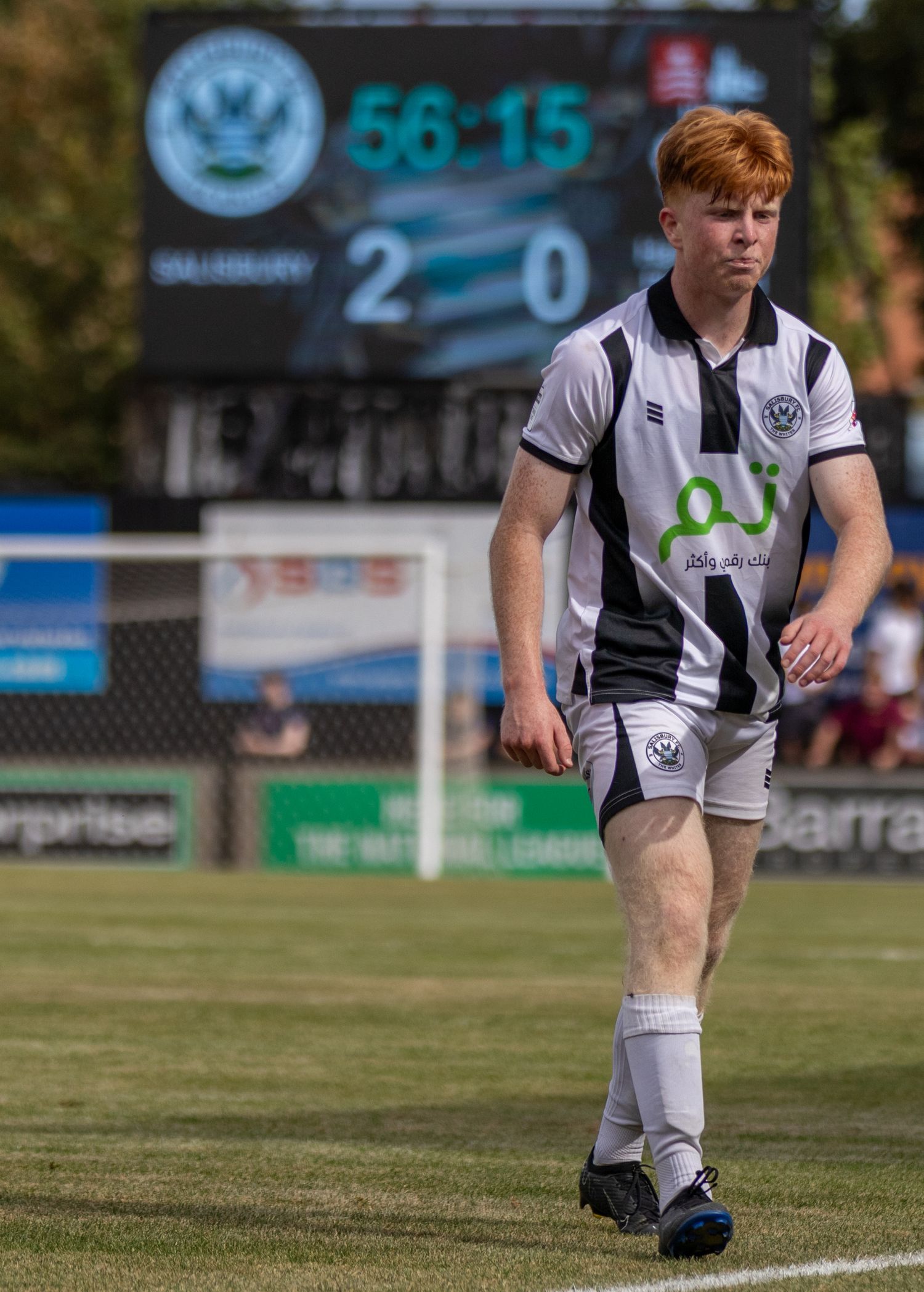 A young red-haired footballer in black and white kit walks on the pitch with a 2-0 scoreboard.