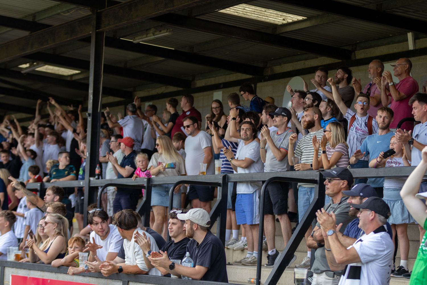Crowd of football supporters clapping and cheering in a covered grandstand during a sunny game.