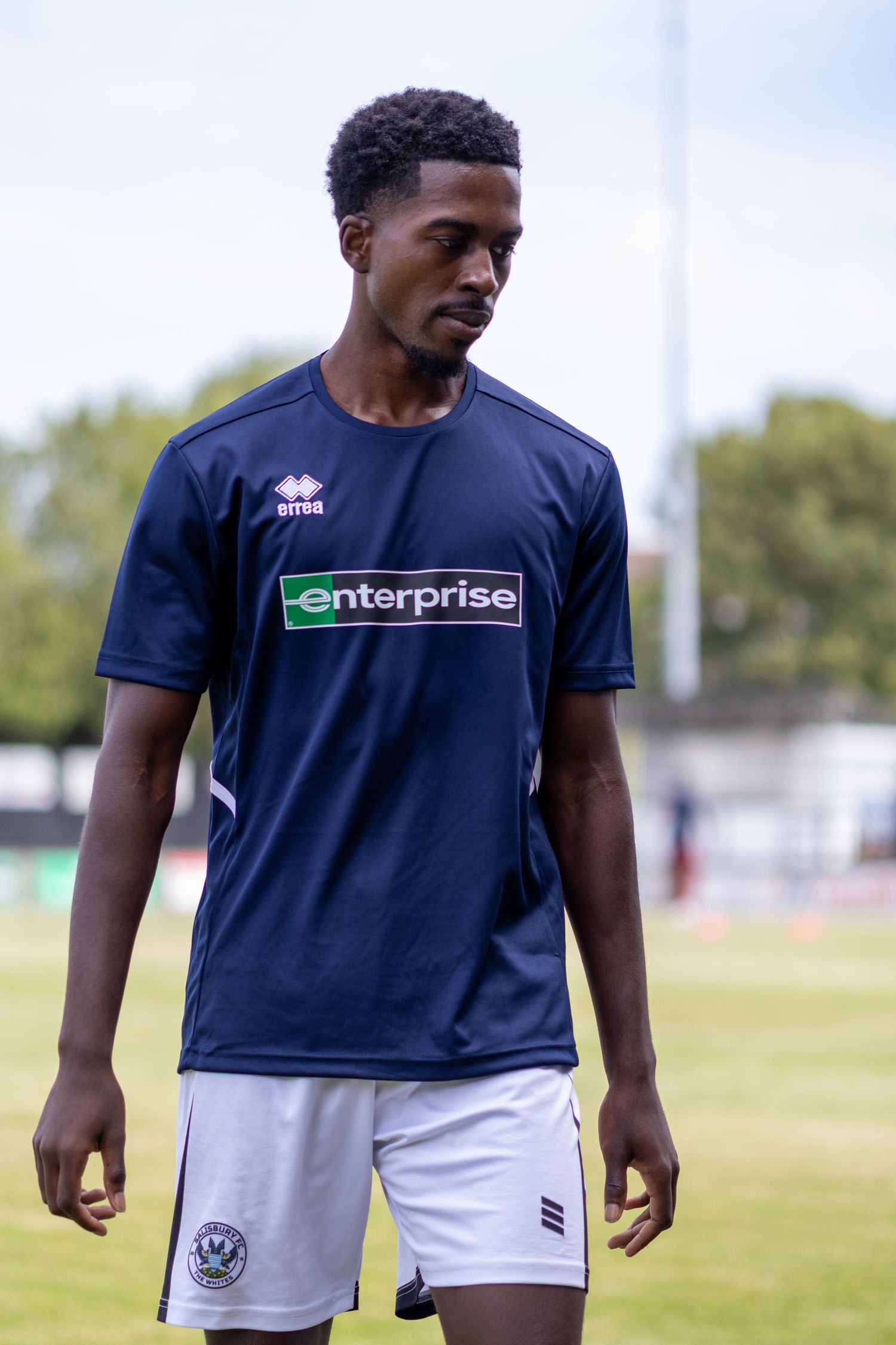 Young Black footballer in navy Enterprise Errea kit standing on football pitch during warmup.