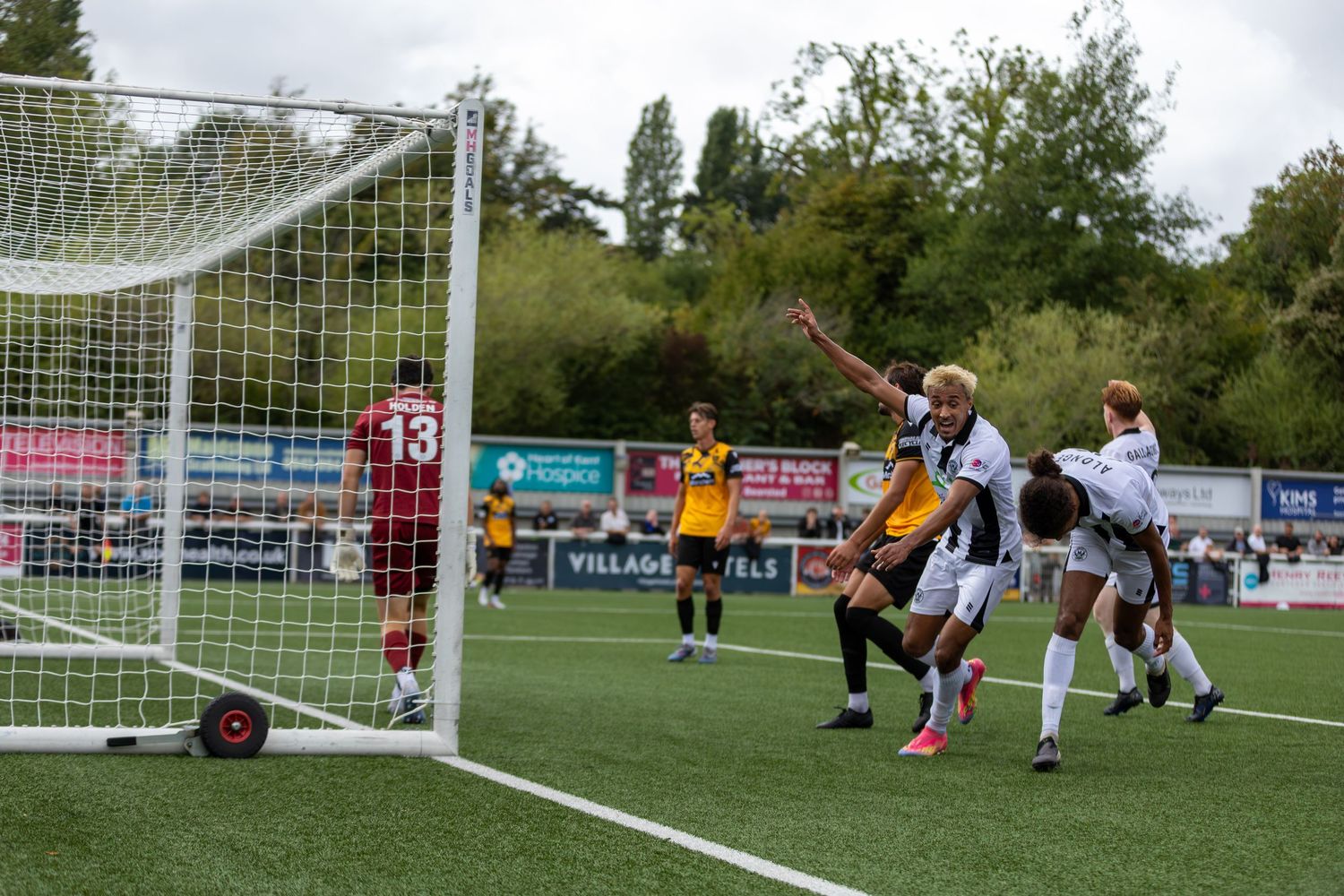 Soccer players celebrate a goal during a match, with goalkeeper in red watching near the net on a green pitch.
