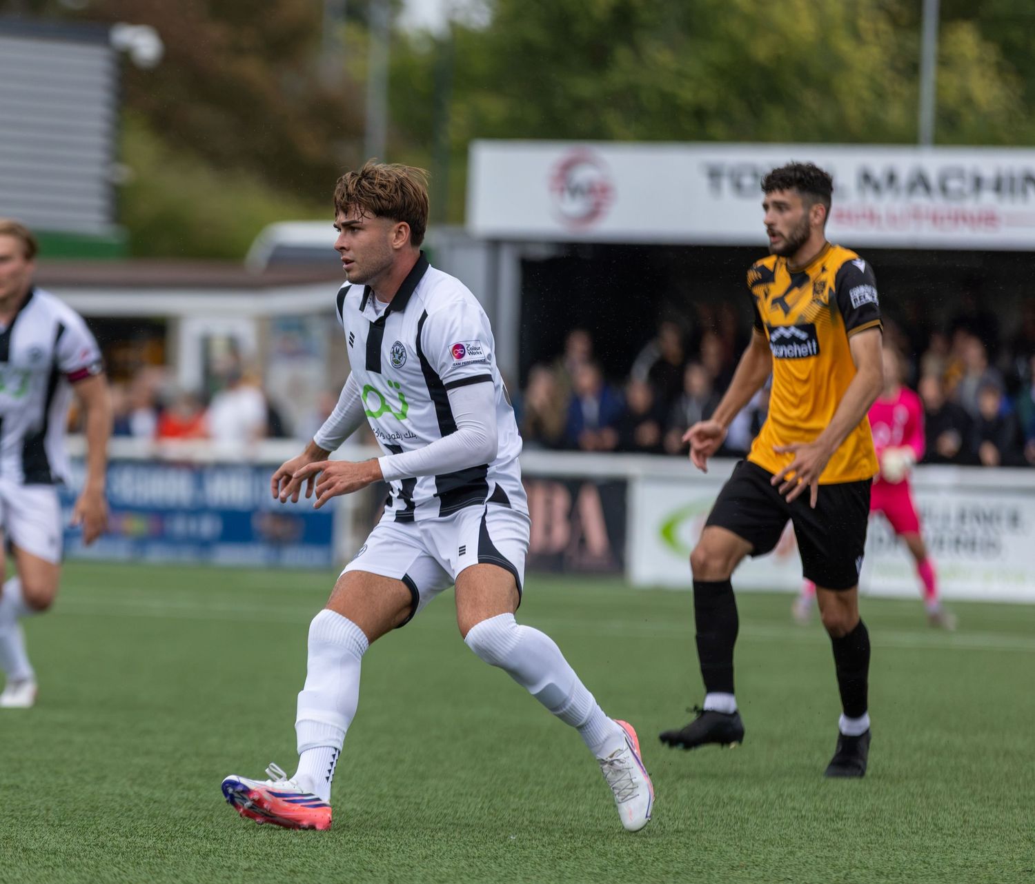 Two soccer players in action during a match, one in black and white kit, the other in yellow, with fans in the background.