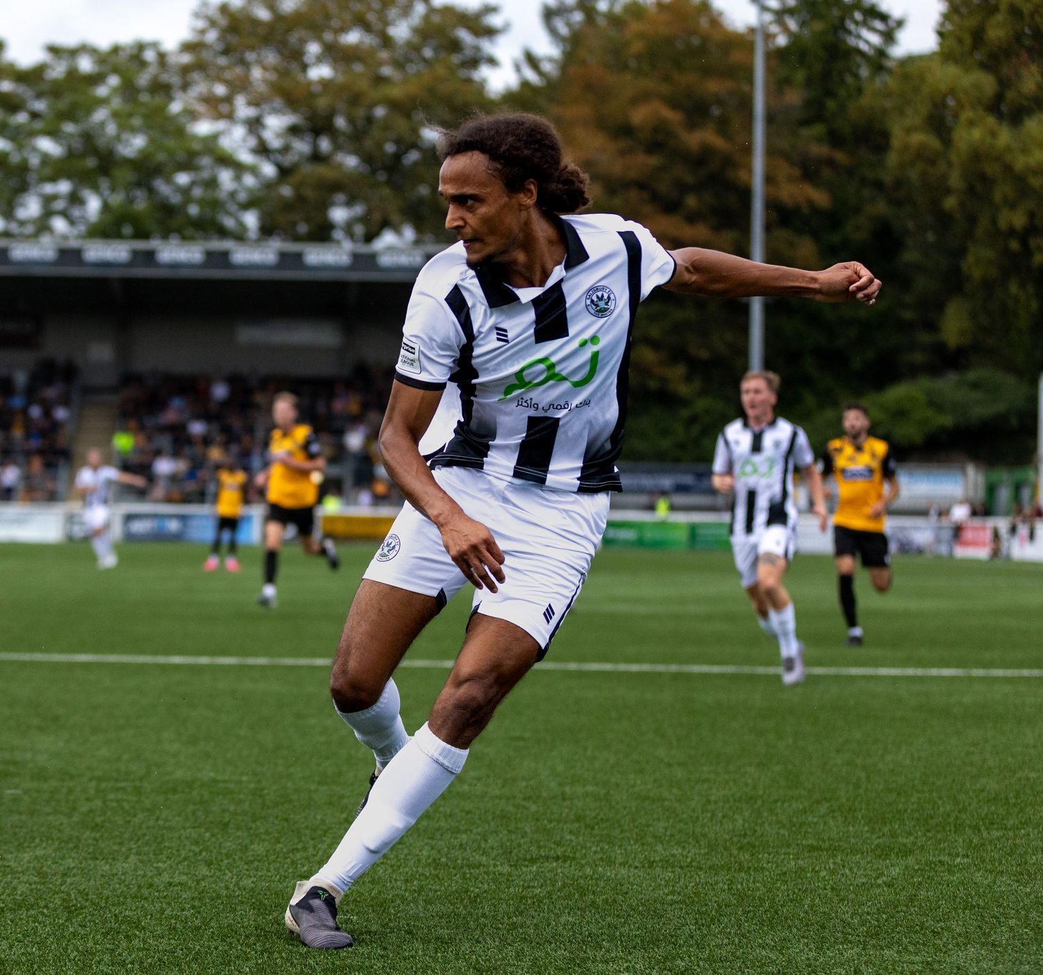 Soccer player in black and white kit dribbling on green pitch during a competitive match with fans in background.