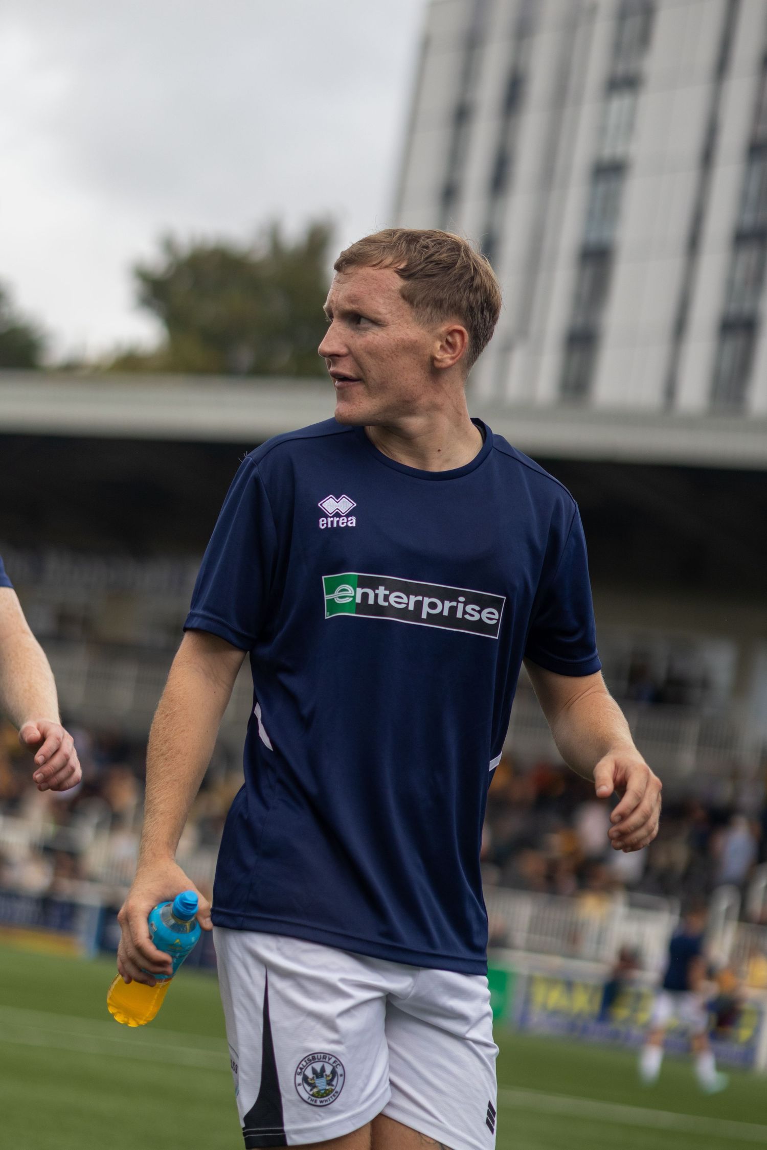 Football player in navy Enterprise-sponsored jersey holding water bottle on pitch.