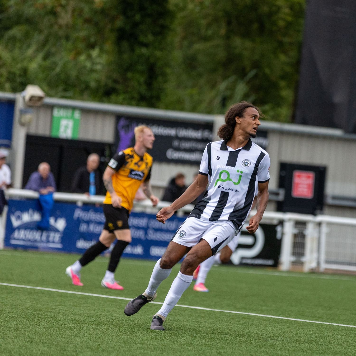 Football player in black and white striped kit celebrates during a match on an artificial pitch.