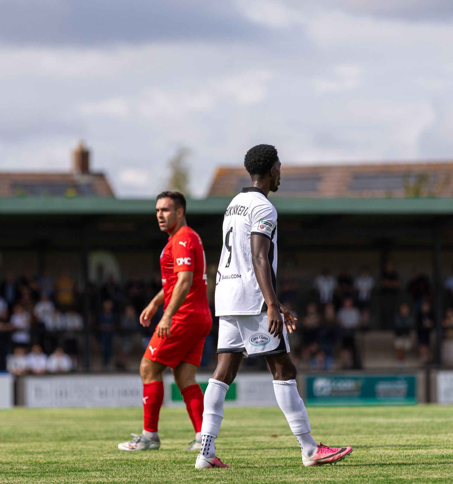 Two soccer players, one in red and one in white, walk on a green pitch during a sunny outdoor match.
